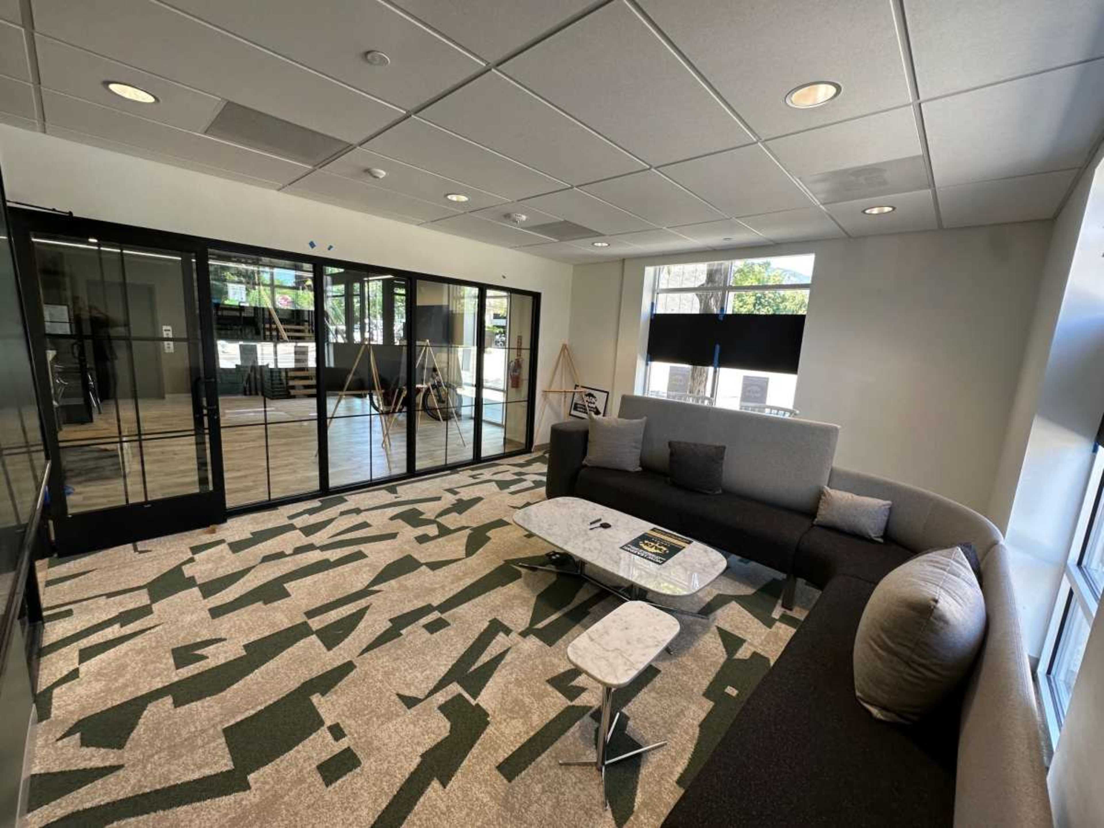 The image shows a modern office lounge area featuring a curved sofa, a minimalist coffee table, and large glass walls that separate it from an adjoining space.