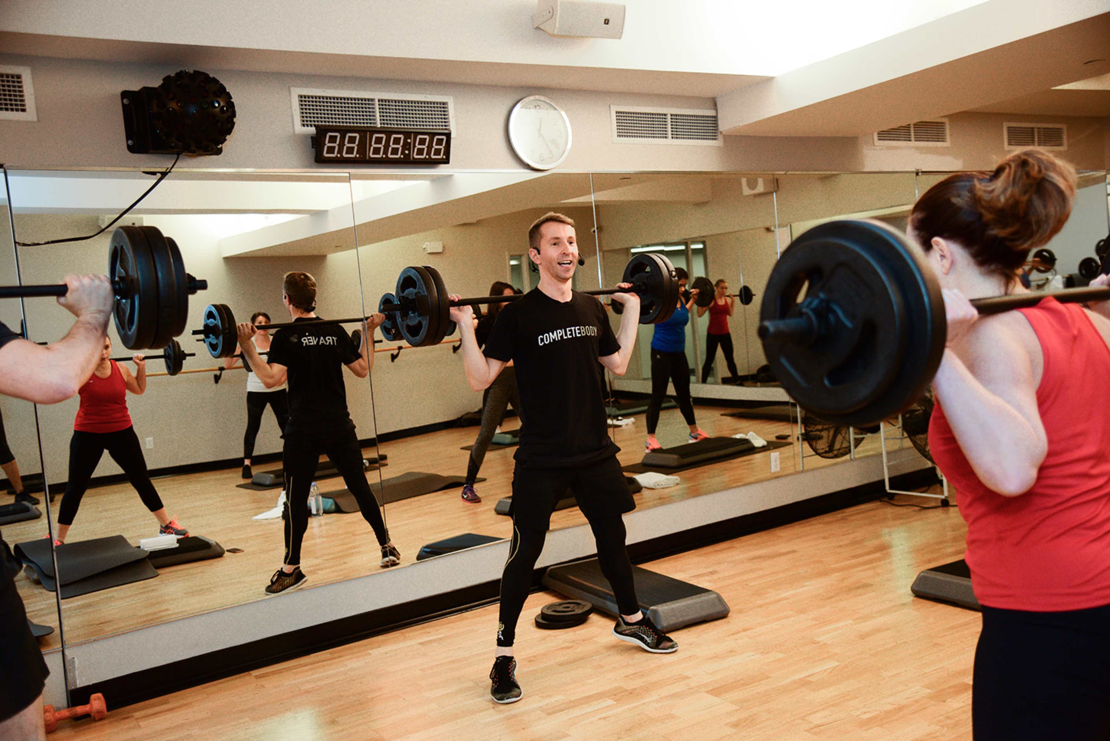 A fitness class is taking place in a gym, with participants lifting barbells while facing a mirror.