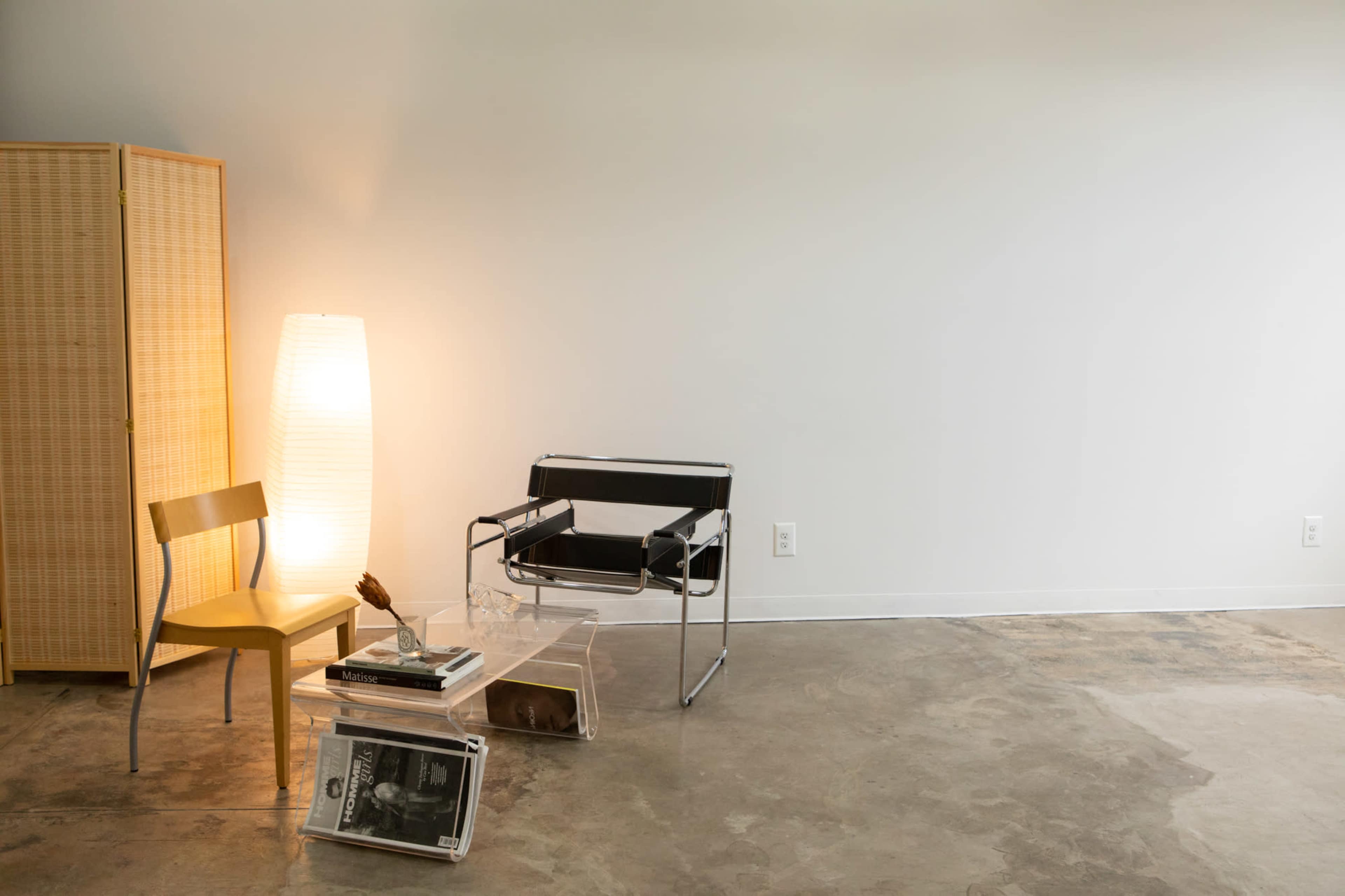 A minimalist interior with a black and chrome chair, a wooden chair, a tall lamp, and a small side table stacked with magazines, set against a light-colored wall.