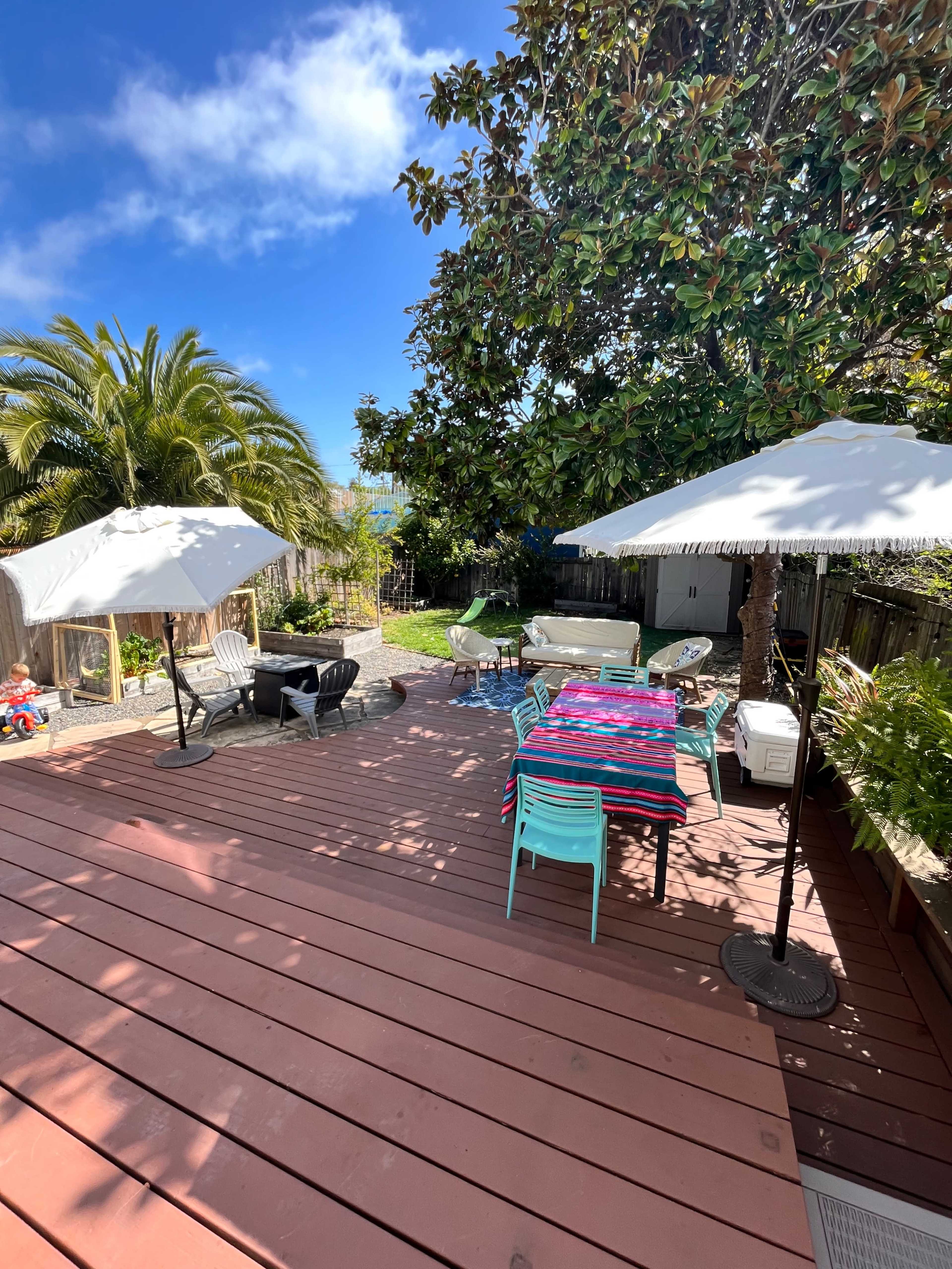 A wooden deck features patio furniture, colorful table settings, and umbrellas, with greenery and a pathway leading to a garden in the background.