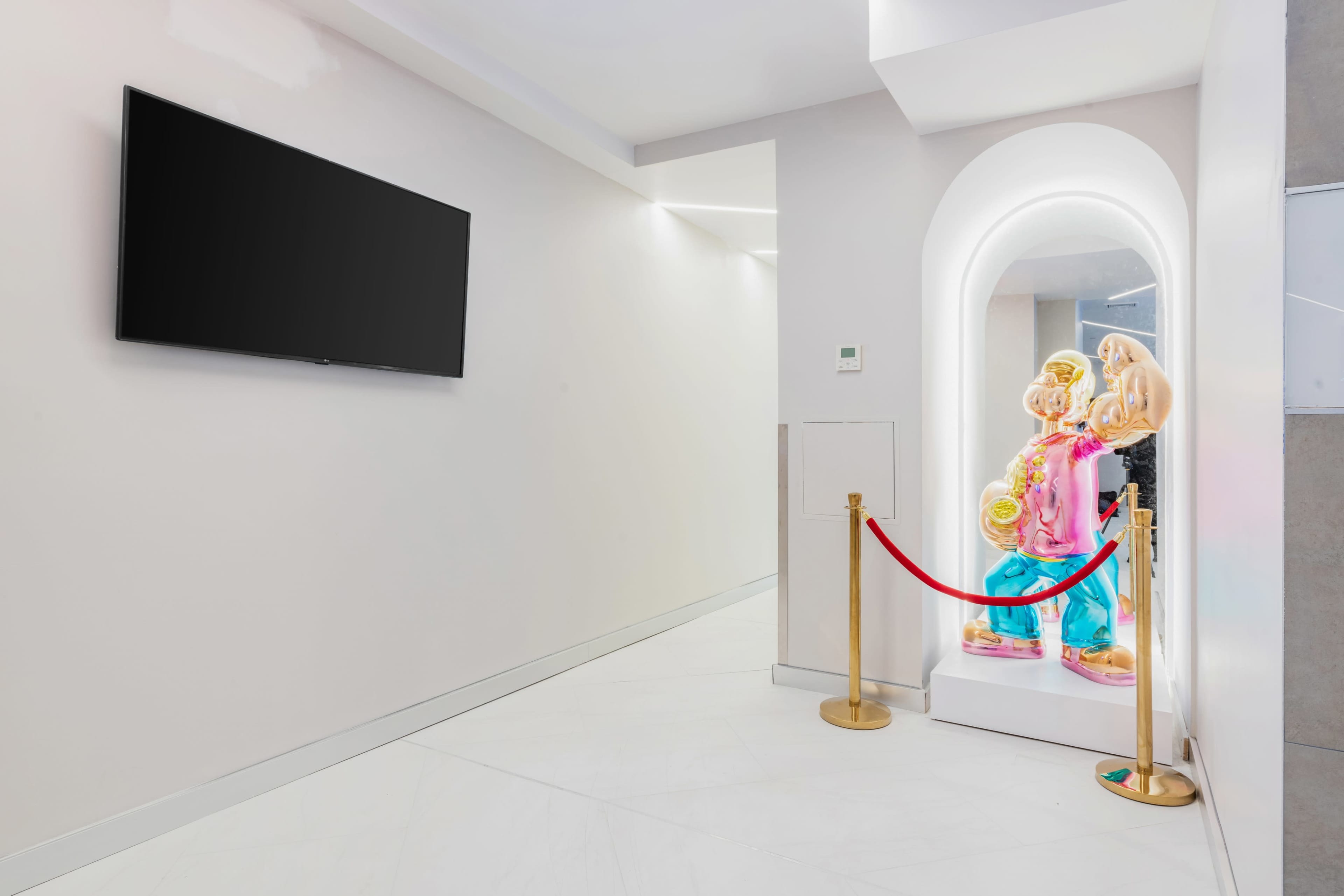 A brightly lit corner of a room features a television mounted on the wall and a colorful statue protected by a red rope barrier.