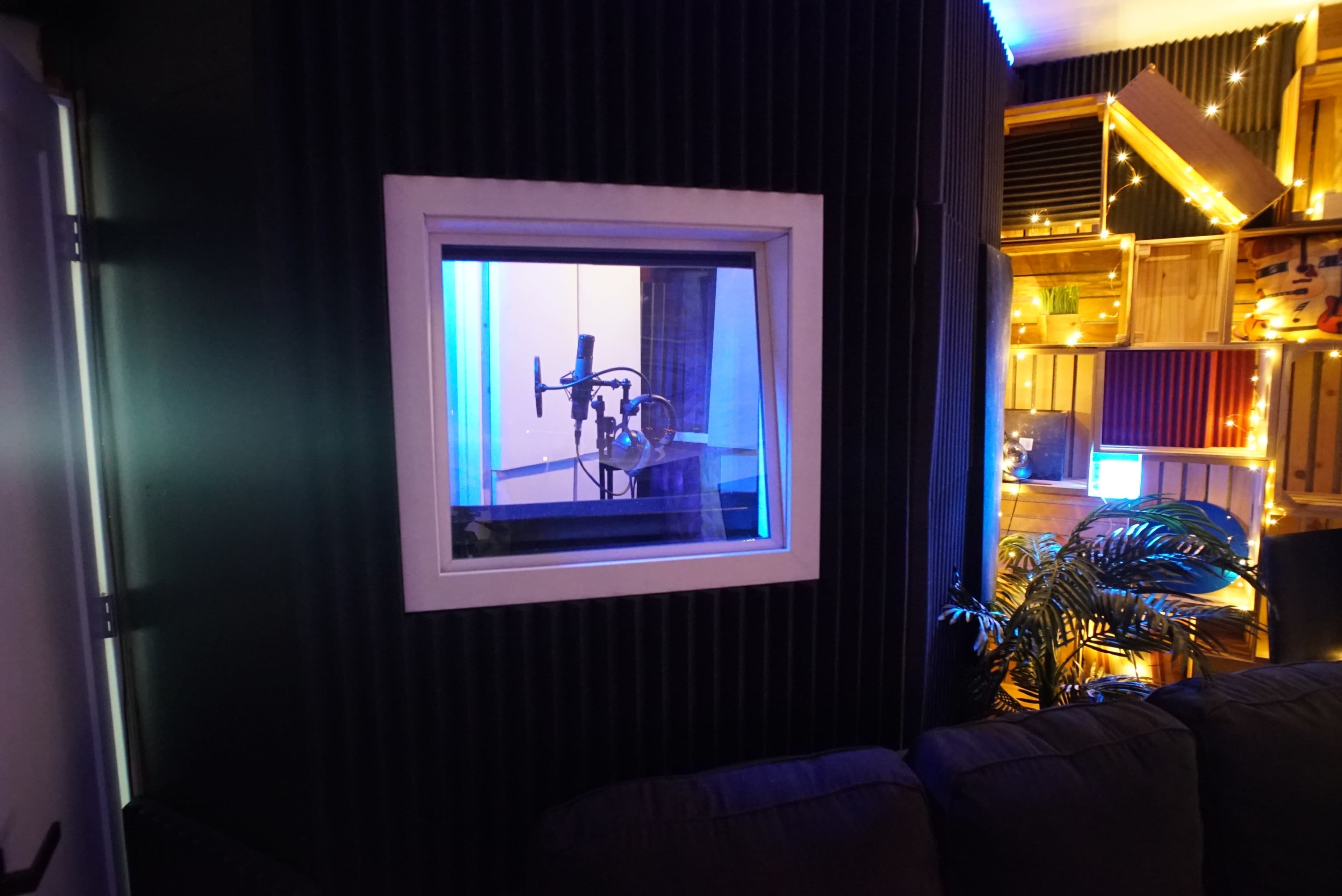 A recording studio with a microphone positioned behind a soundproof glass window, illuminated by blue lighting.