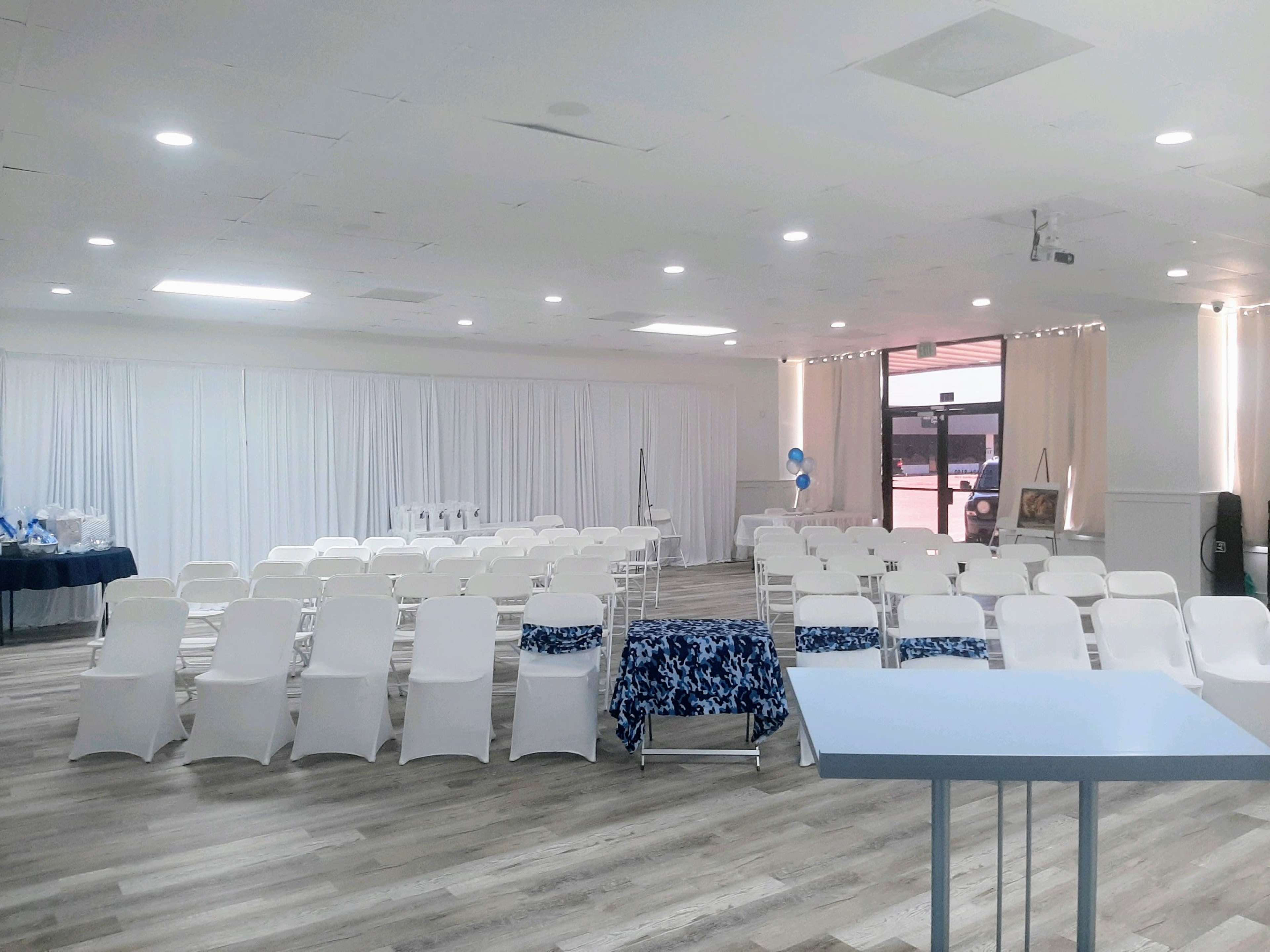 A spacious event room set up with rows of white chairs facing a blank podium, with blue and white decorations and drapes along the walls.