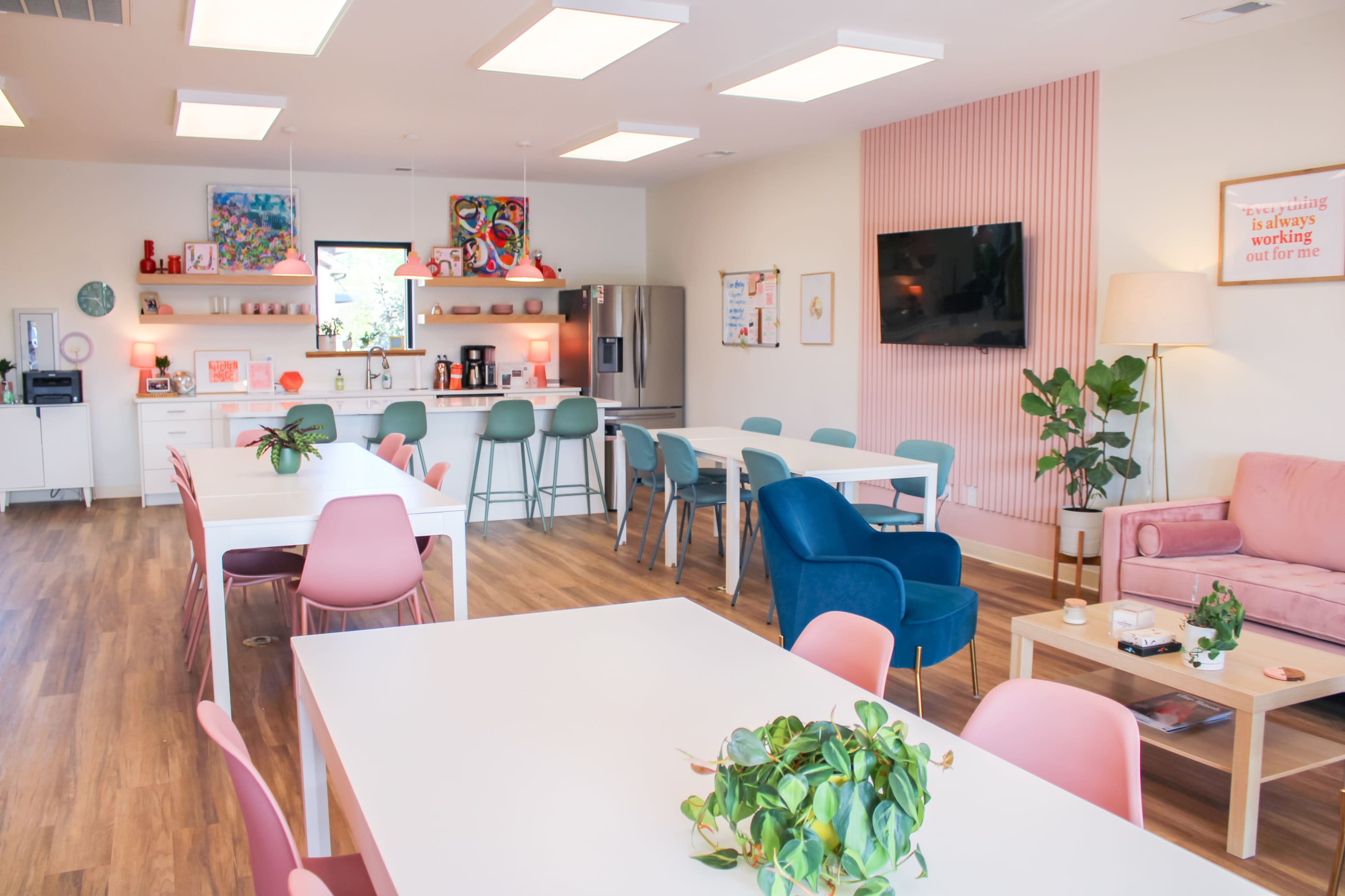 A brightly lit communal workspace featuring several tables, a kitchenette, and a lounge area with modern furnishings and indoor plants.