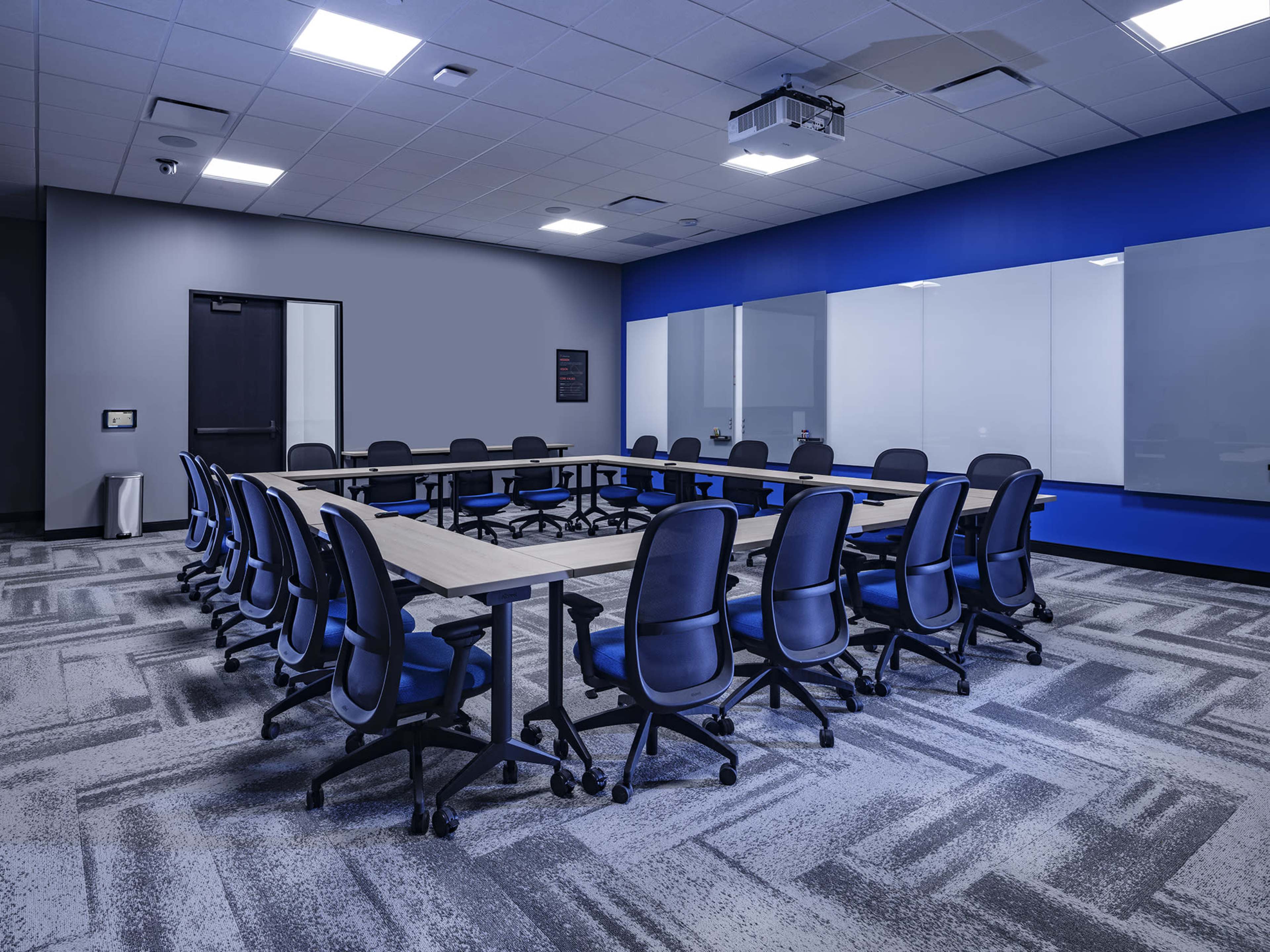 A modern conference room features a large rectangular table surrounded by ergonomic office chairs, with a projection screen and whiteboards on the walls.