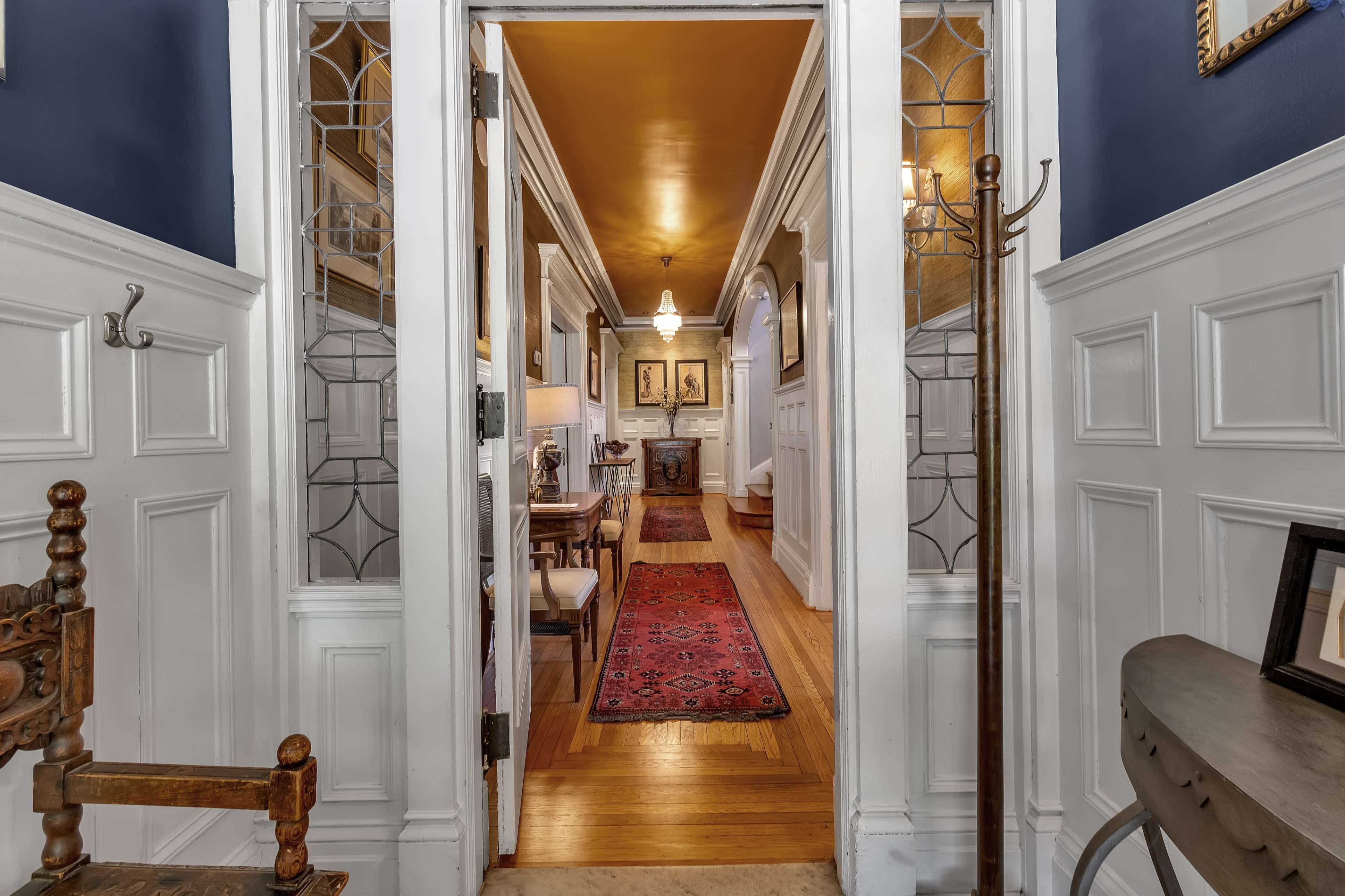 A hallway with white wainscoting and blue walls, leading to a brightly lit room adorned with a red rug and framed artwork.