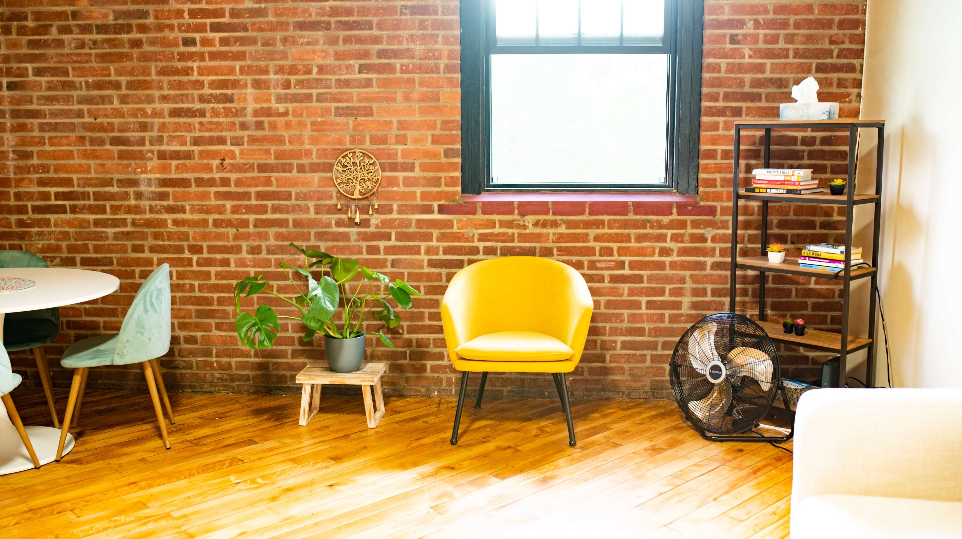 A bright, minimalist living space with a yellow chair, a potted plant, a bookshelf, and a window along a brick wall.