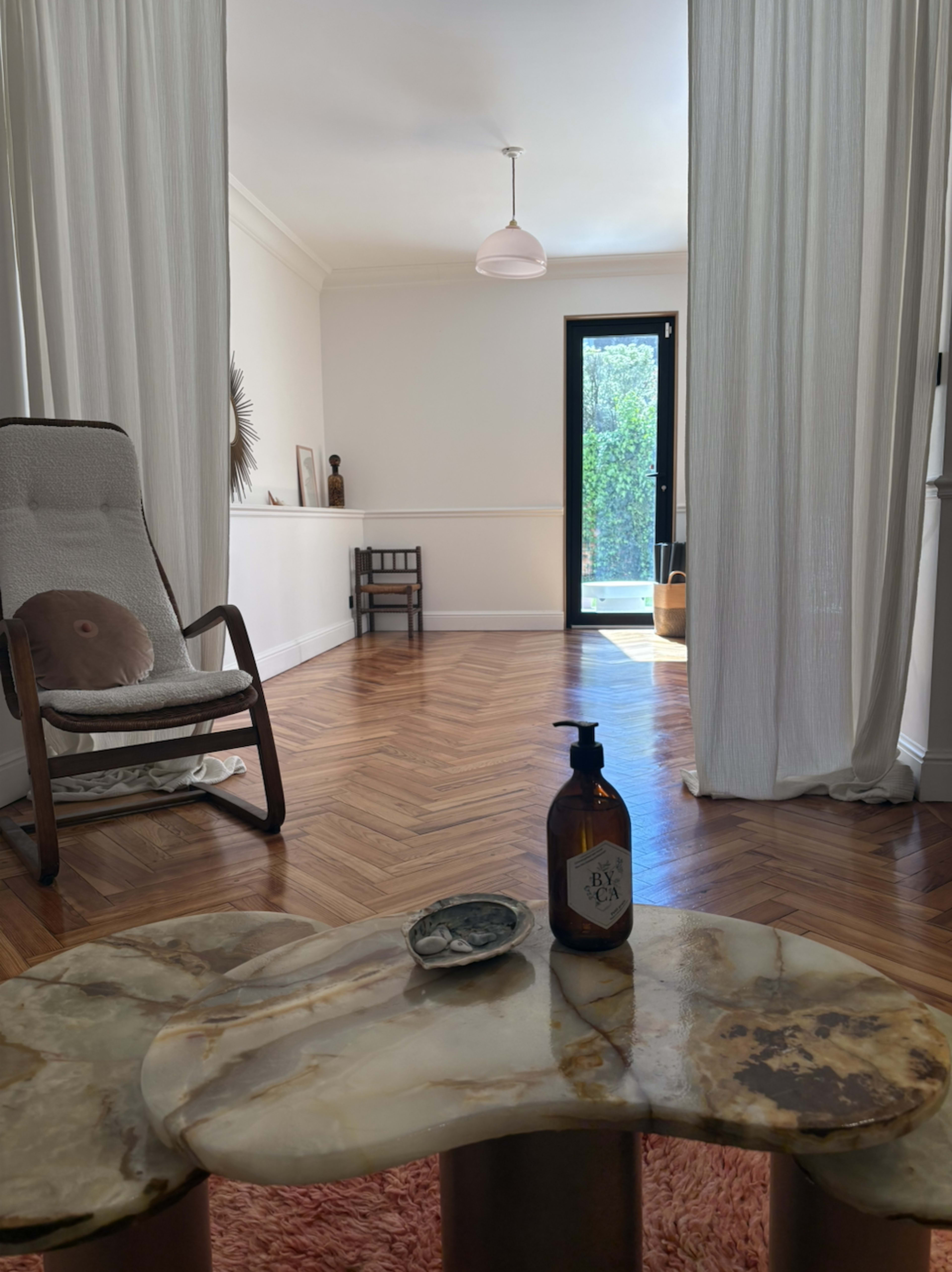 A well-lit room features a rocking chair and a small table with a bottle and dish, framed by sheer curtains and an open door leading to an outdoor space.