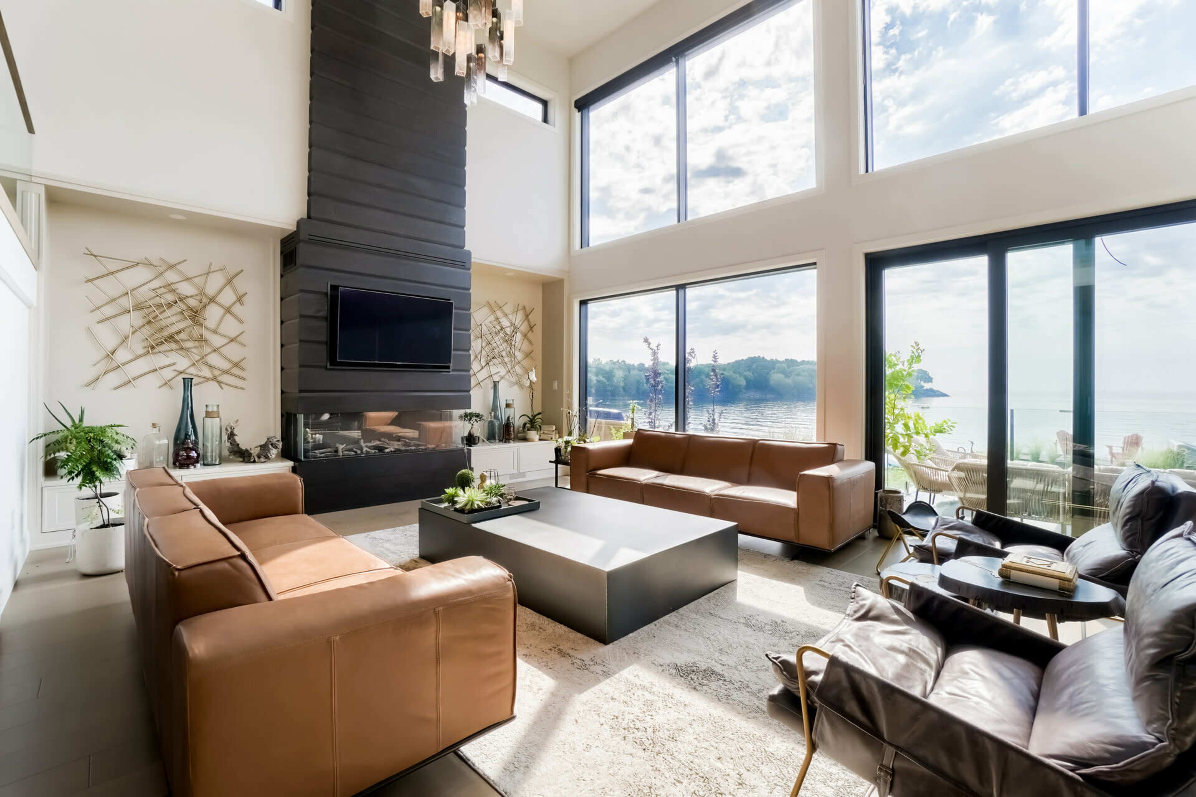 A modern living room with large windows overlooking a lake, featuring brown leather sofas arranged around a rectangular coffee table and a minimalist decor.
