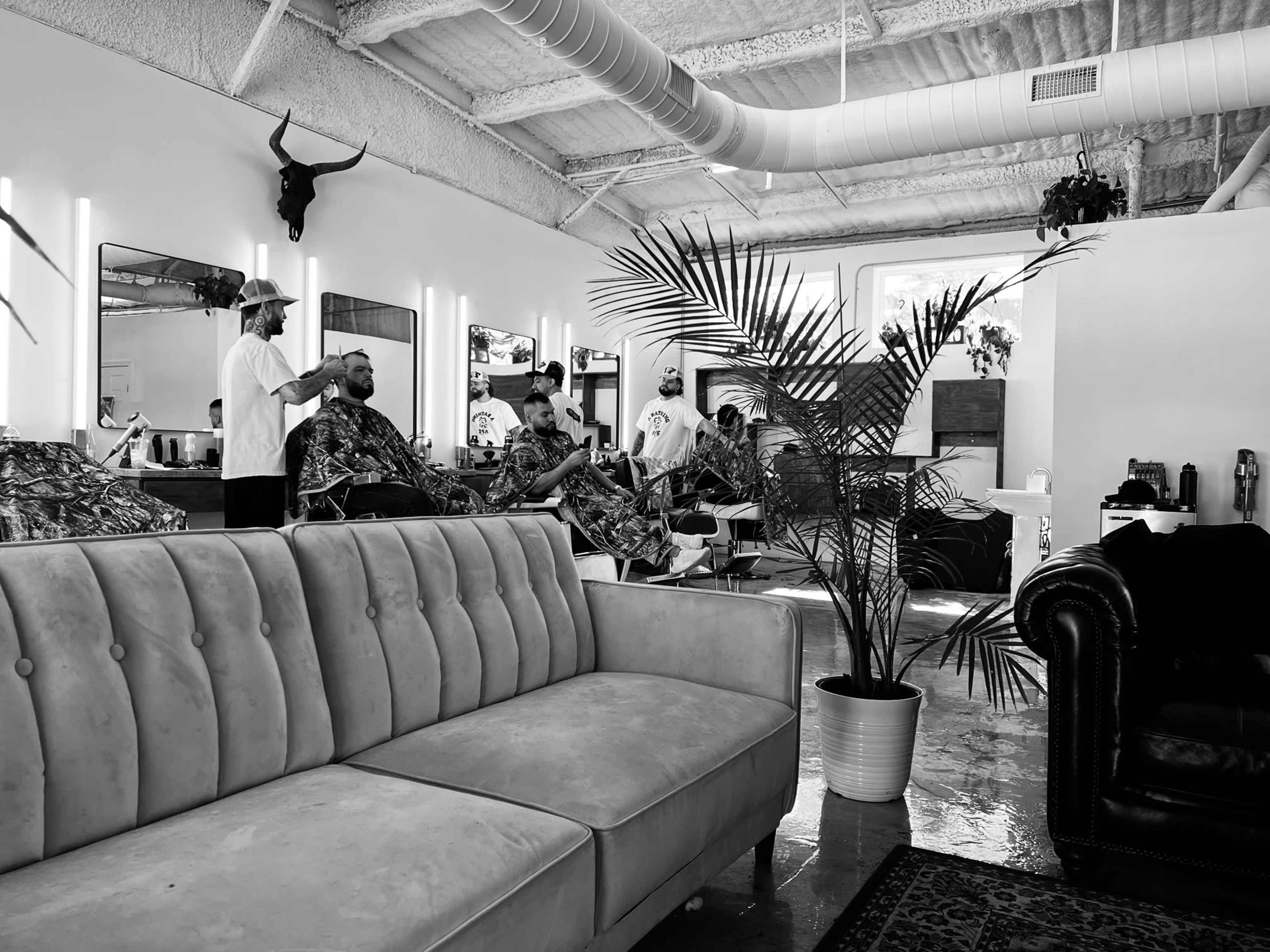 A modern barber shop features stylists attending to customers seated in chairs, with a couch and potted plants visible in the foreground.