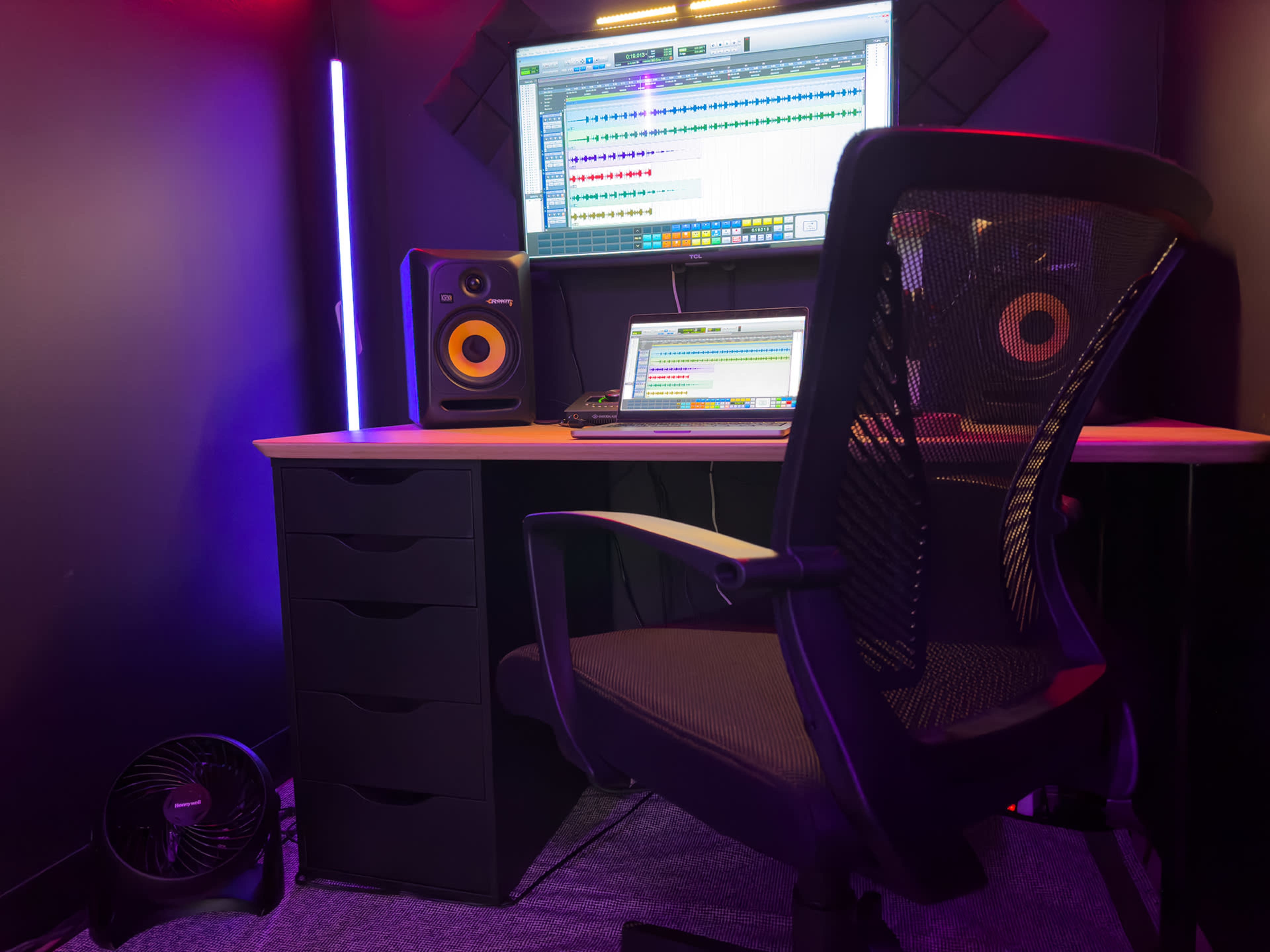 The image shows a recording studio corner with a computer, audio software displayed on the screen, studio monitors, an office chair, and a small fan.