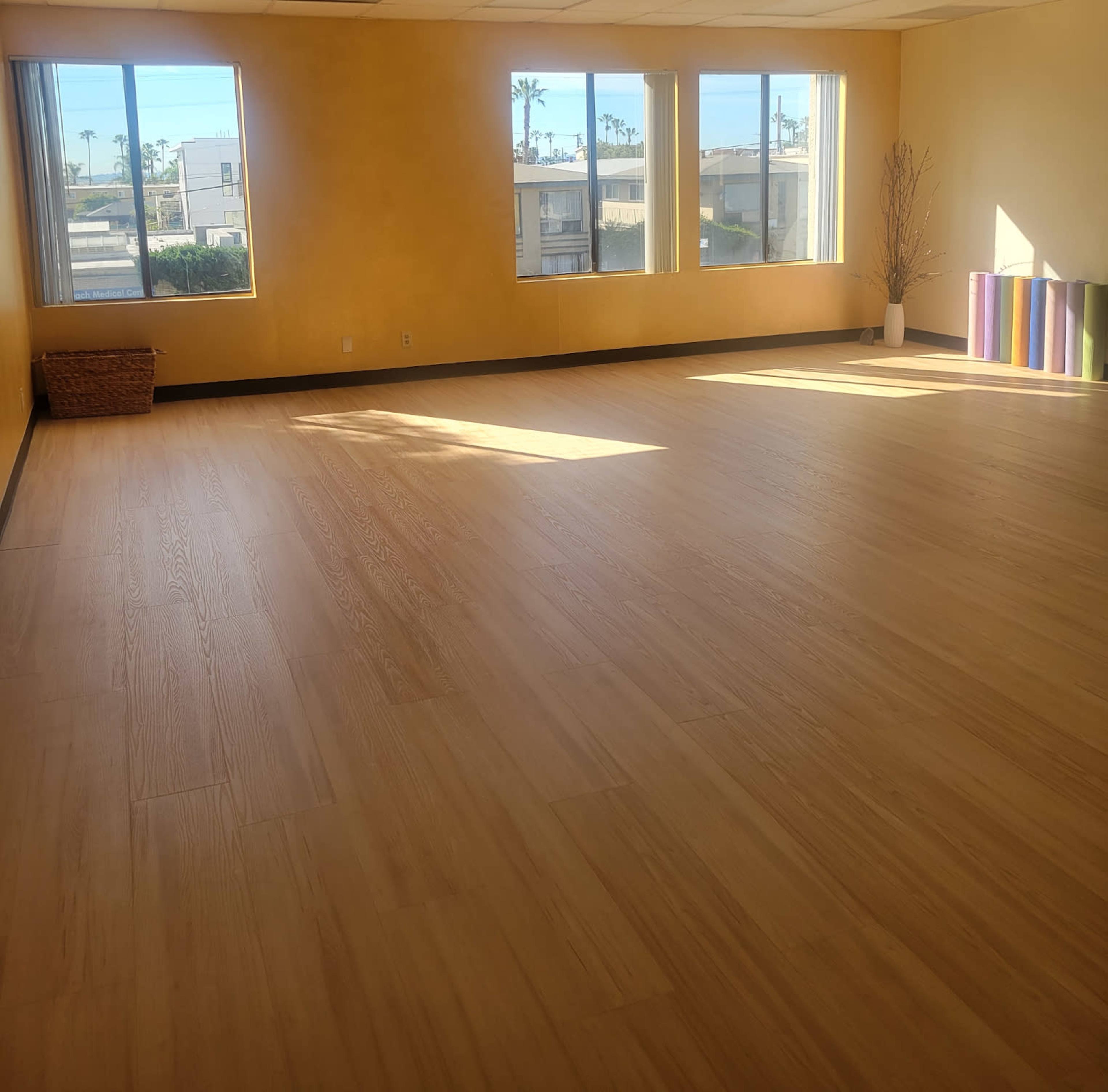 The image shows a spacious, empty room with large windows and light-colored wooden flooring.