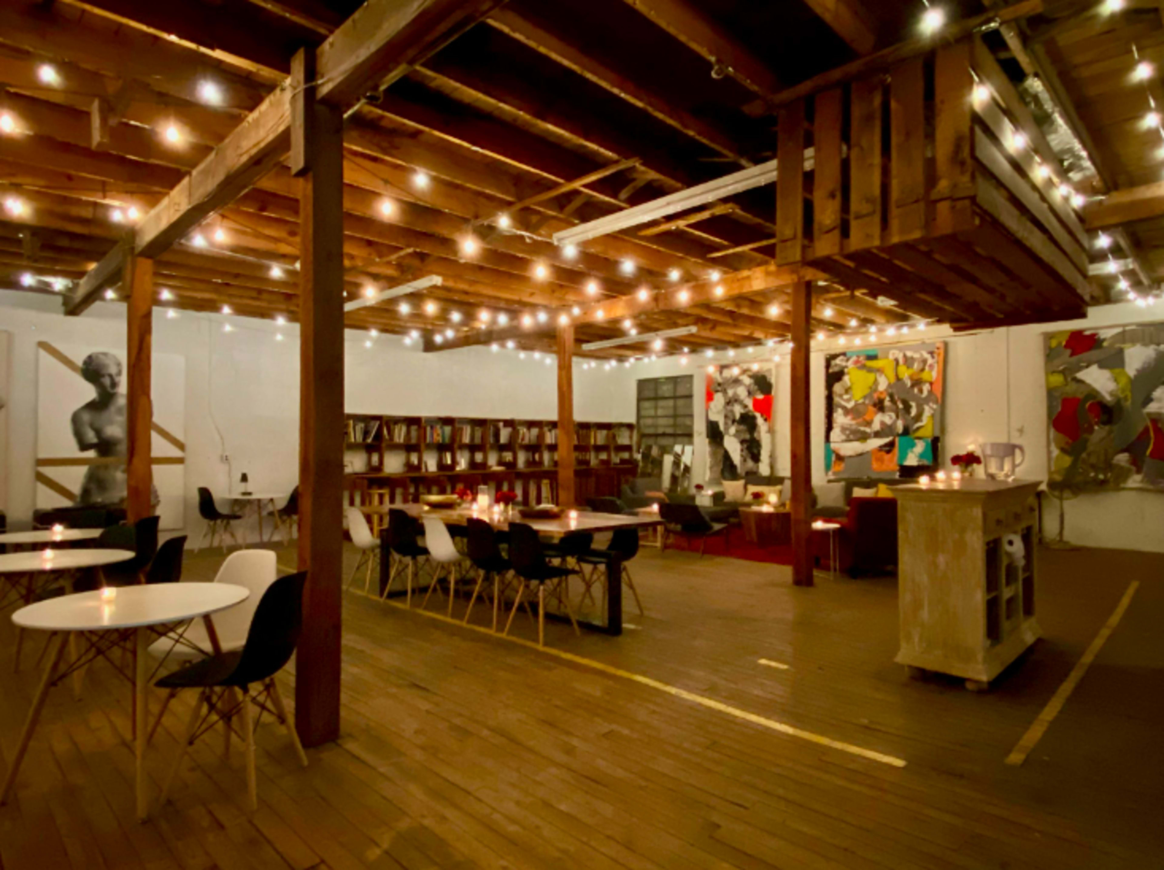 A spacious, well-lit room with wooden floors, exposed beams, and string lights, featuring a mix of dining tables and chairs, along with artwork on the walls.