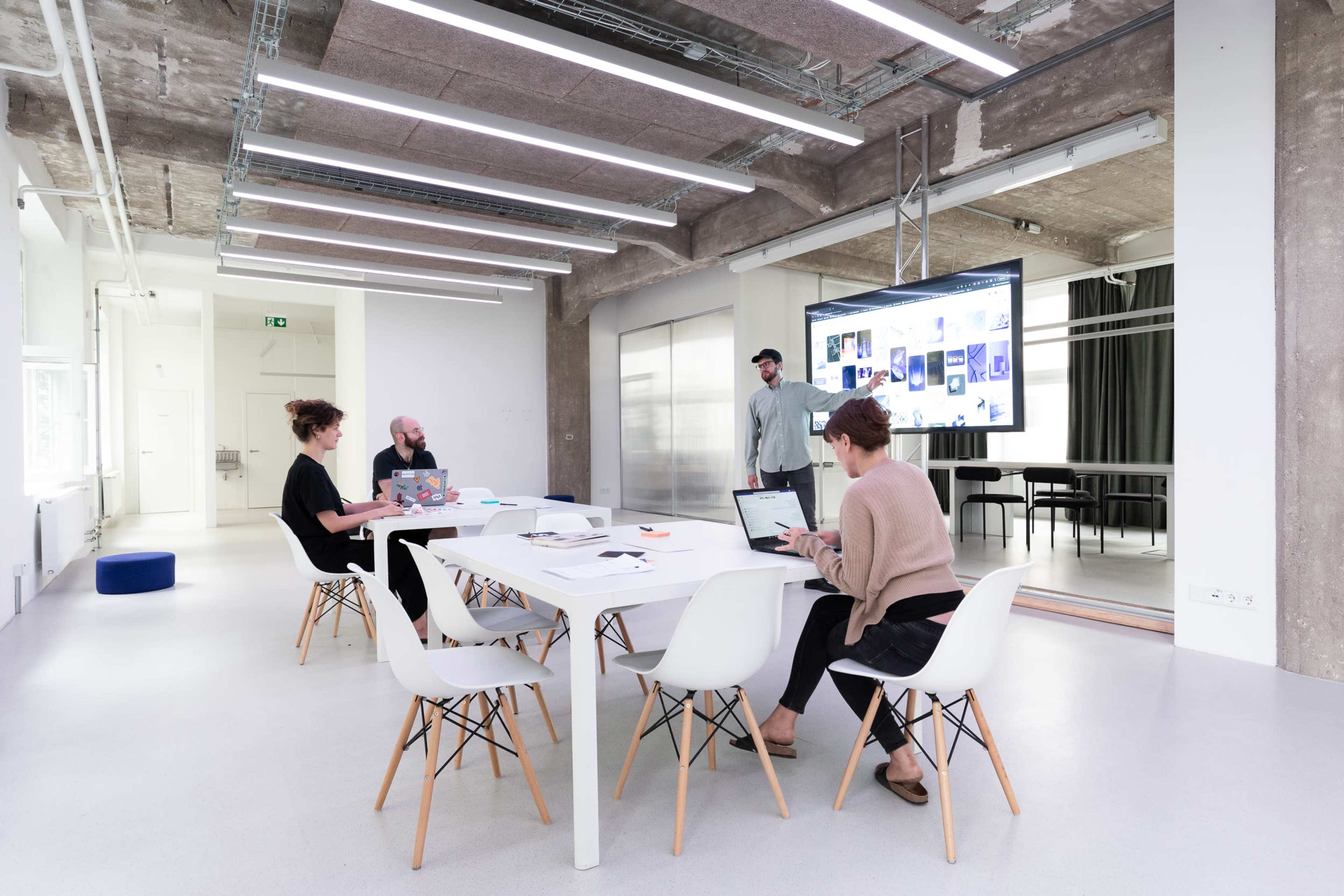 A group of four people work at a modern conference table in a well-lit, minimalist office with a large screen displaying content in the background.