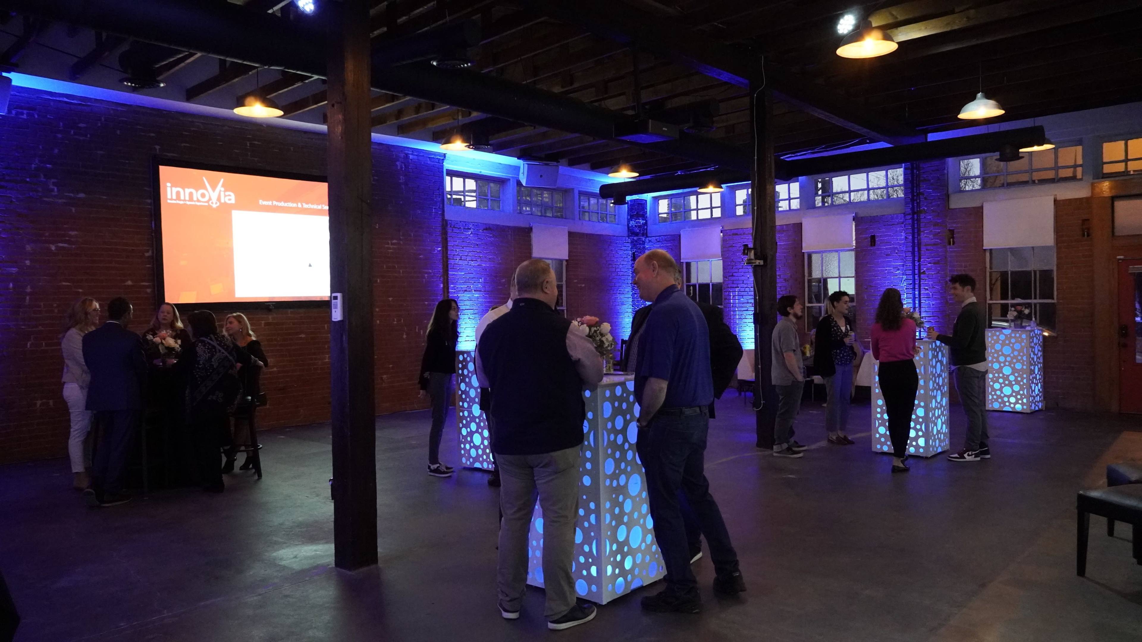 A group of people mingles in a well-lit event space with illuminated tables and exposed brick walls.