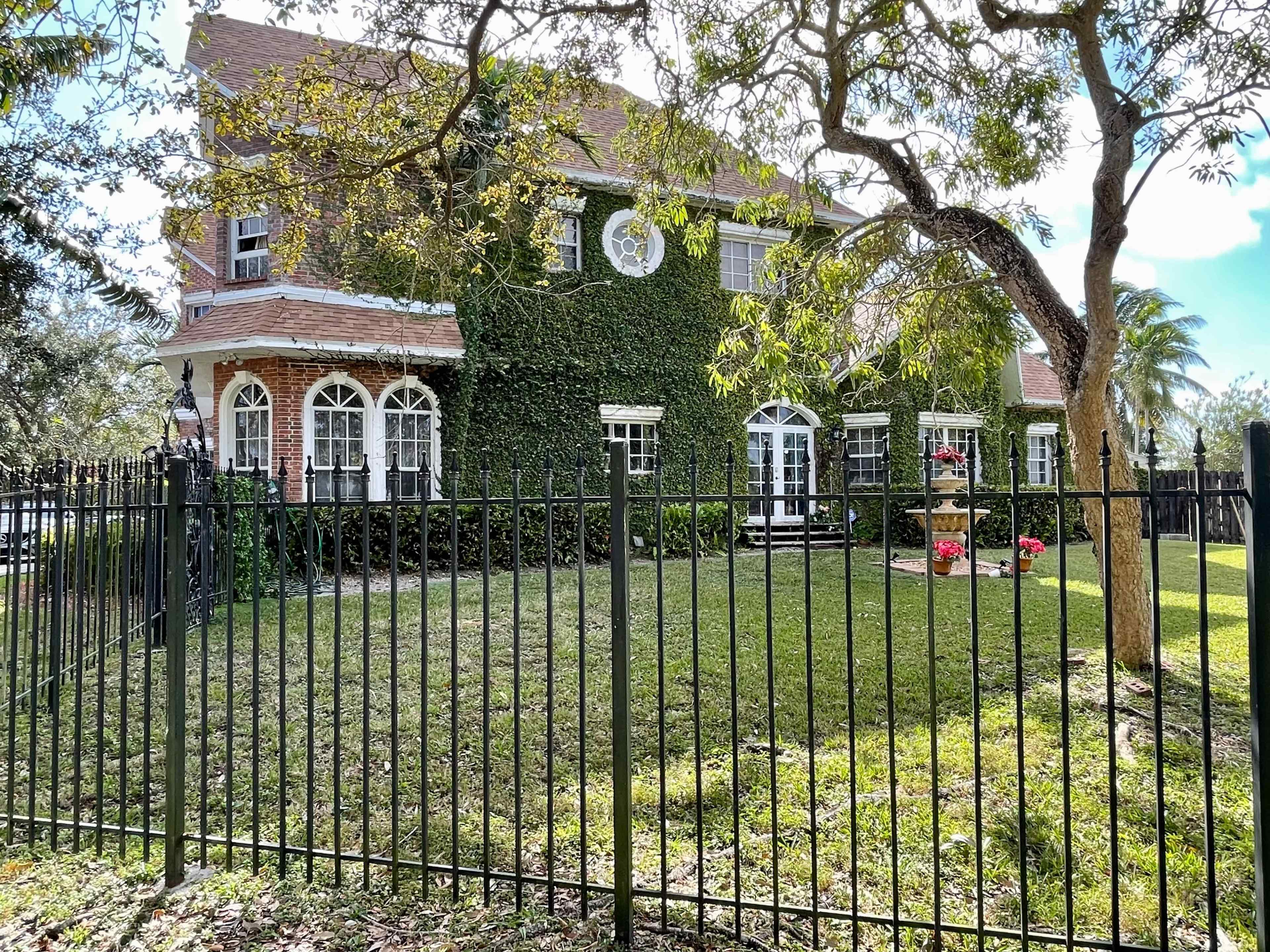 A two-story house covered in greenery is surrounded by a black metal fence and features a well-maintained lawn with a small garden area.