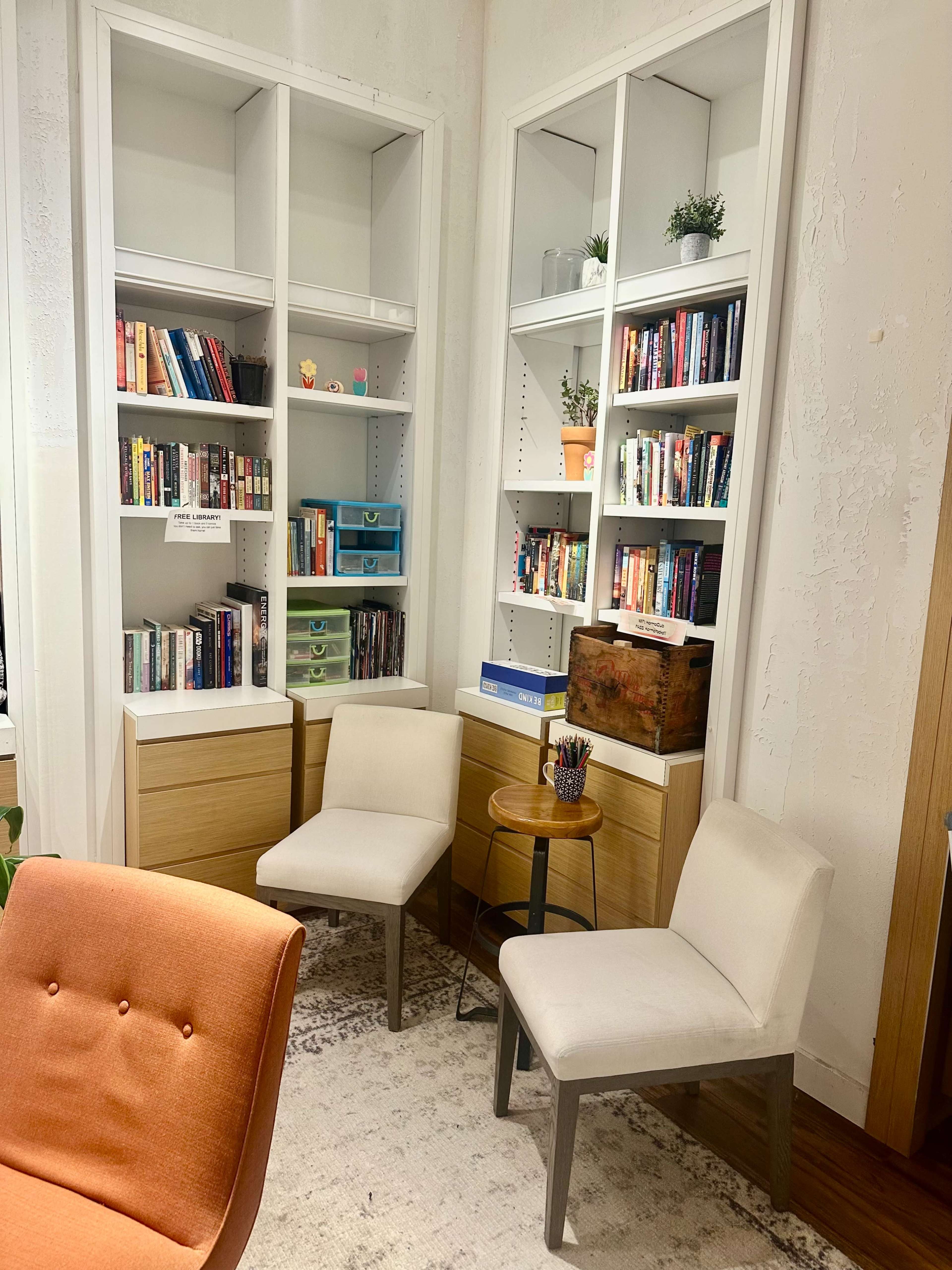 The image shows a corner of a room featuring two white chairs, a small round table, and built-in bookshelves filled with various books and decorative items.
