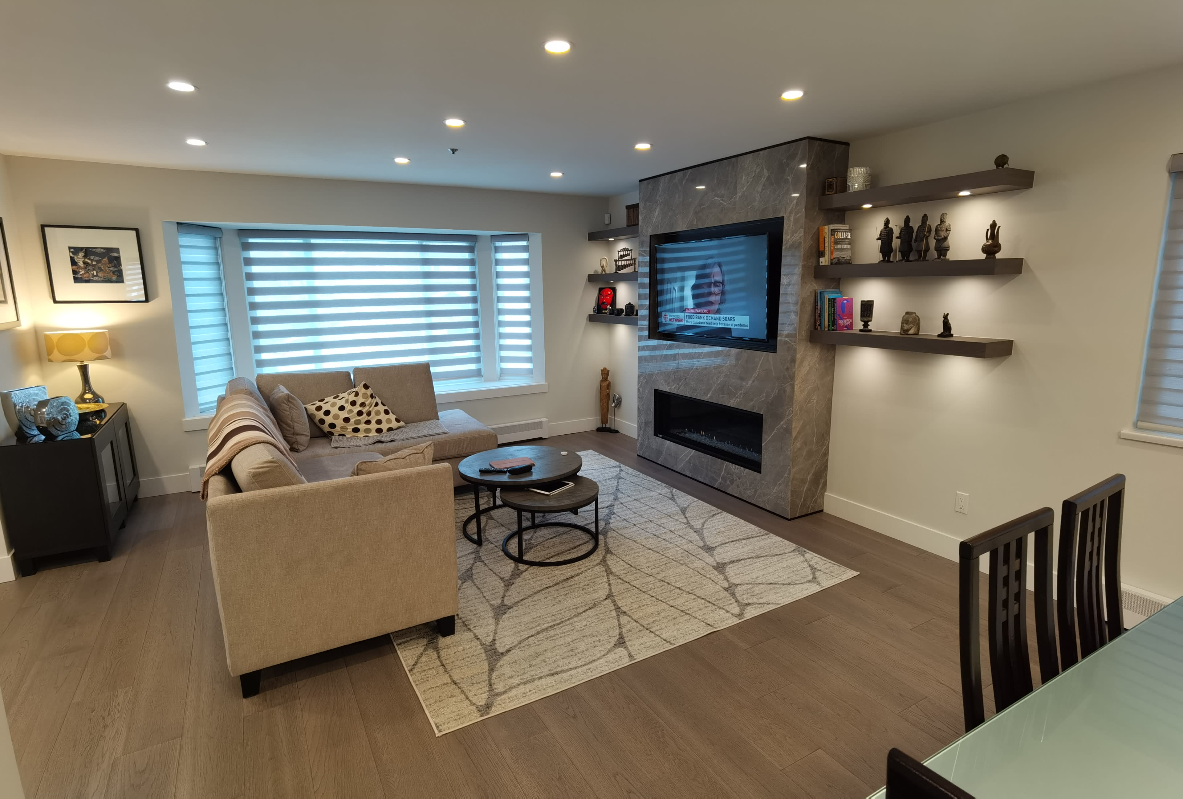 The living room features a sectional sofa, a round coffee table, a wall-mounted TV, and decorative shelves, with soft lighting and a large area rug.