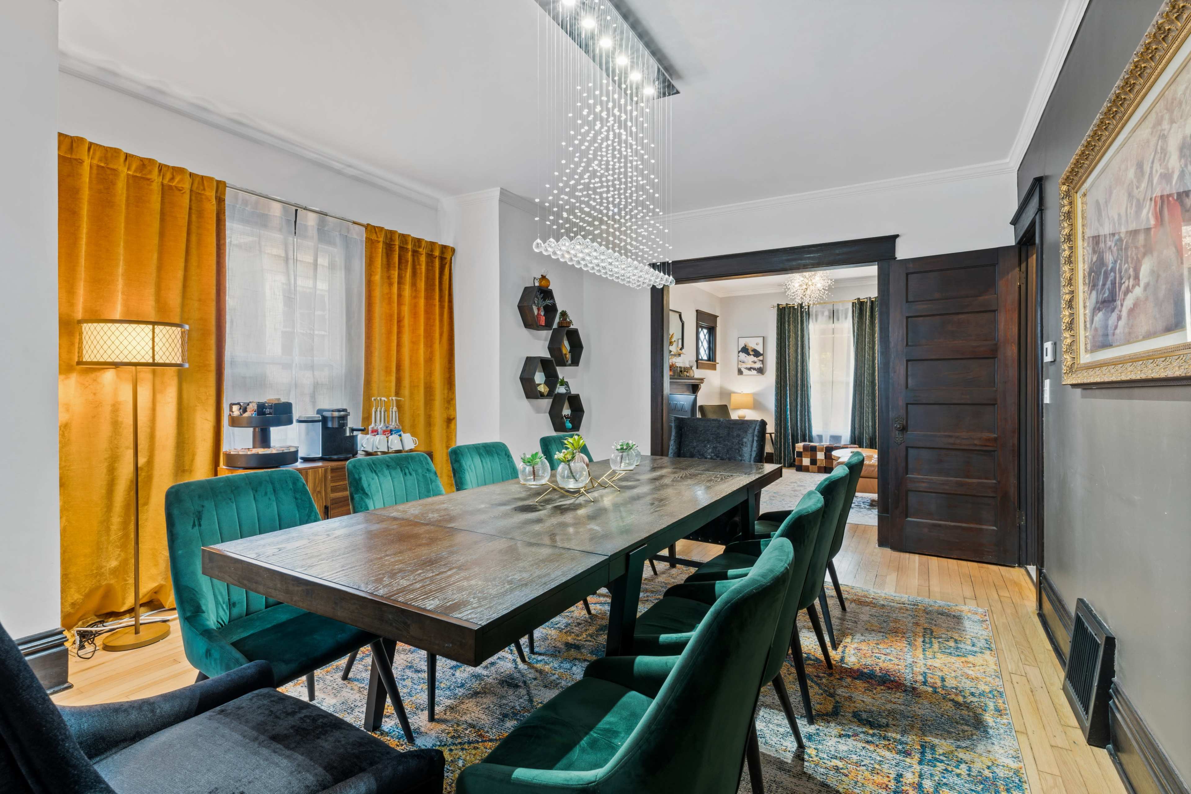The image shows a dining room featuring a long wooden table surrounded by green upholstered chairs, with a modern chandelier overhead and yellow curtains framing a window.