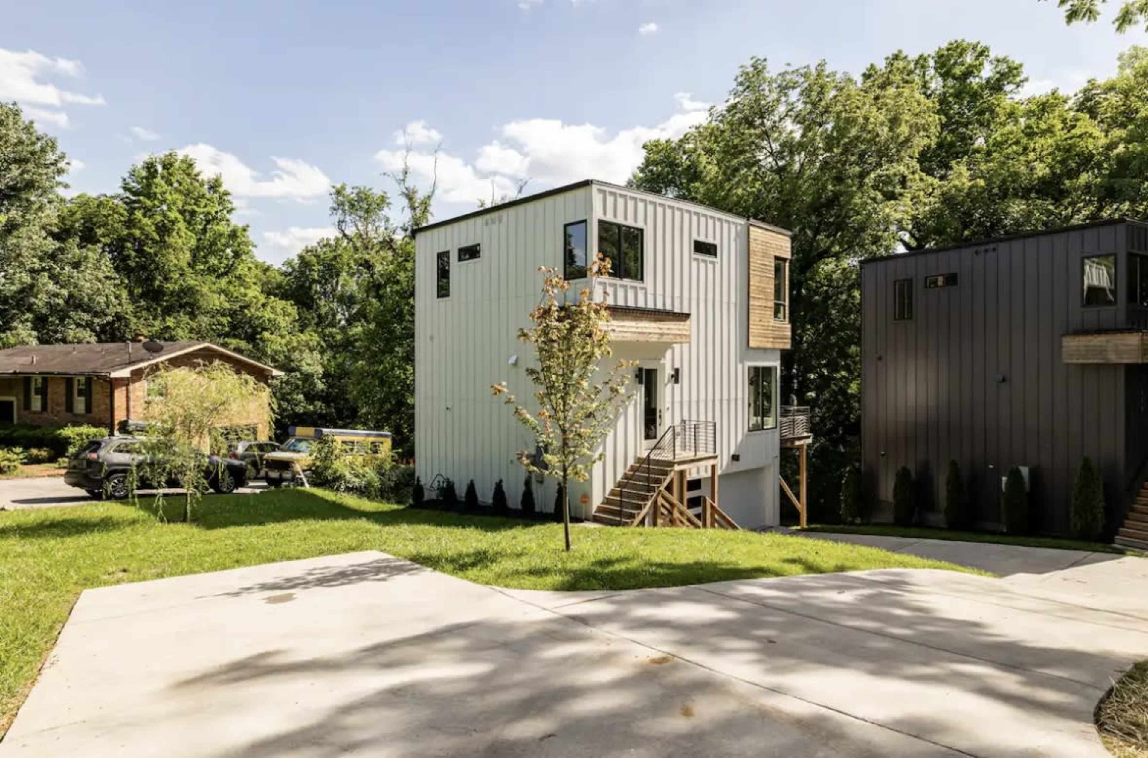 The image shows two modern, multi-story houses set among green trees on a residential street.