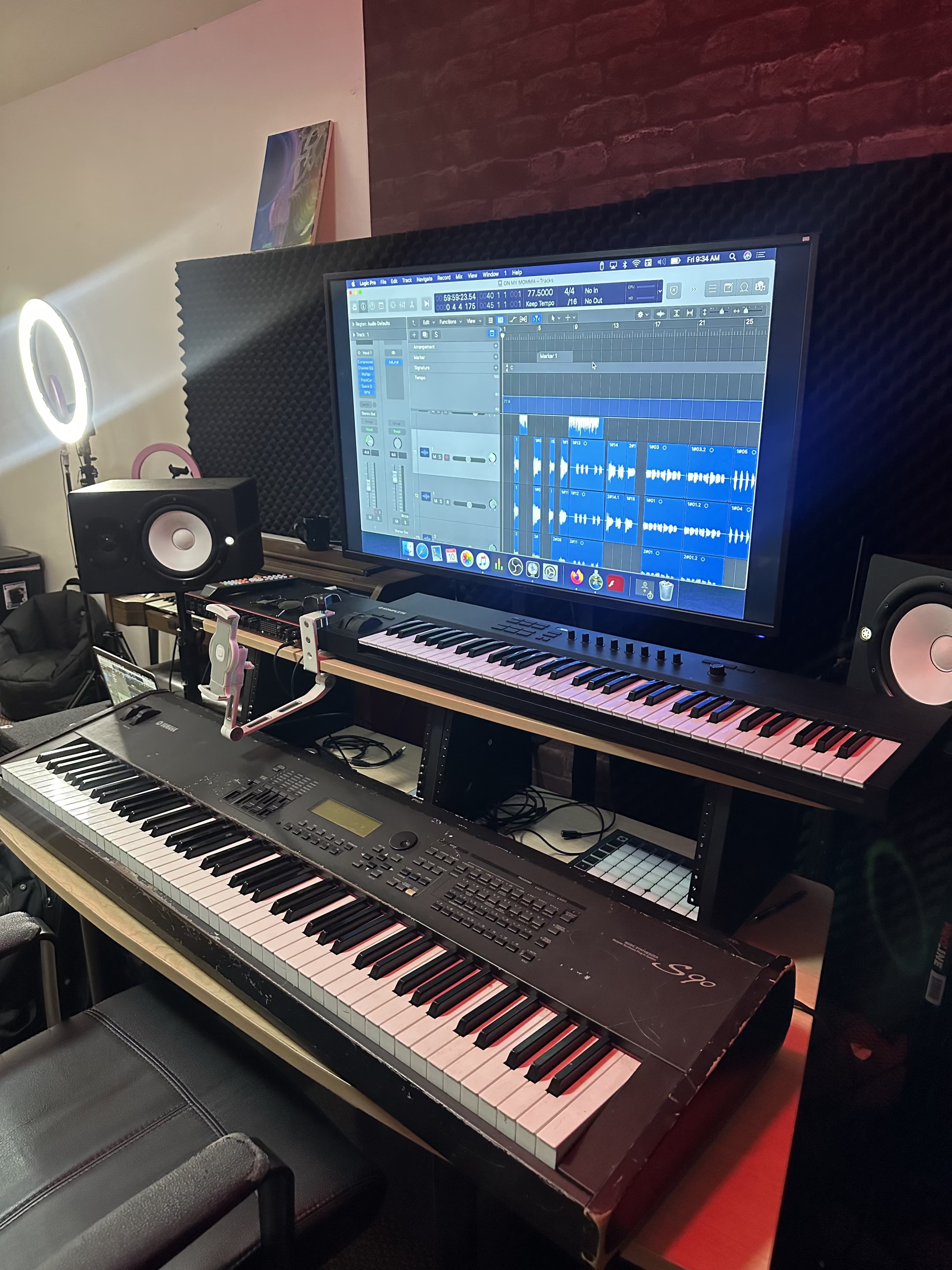 A music production setup features a digital audio workstation displayed on a large monitor, with two keyboards and studio monitors on a desk.