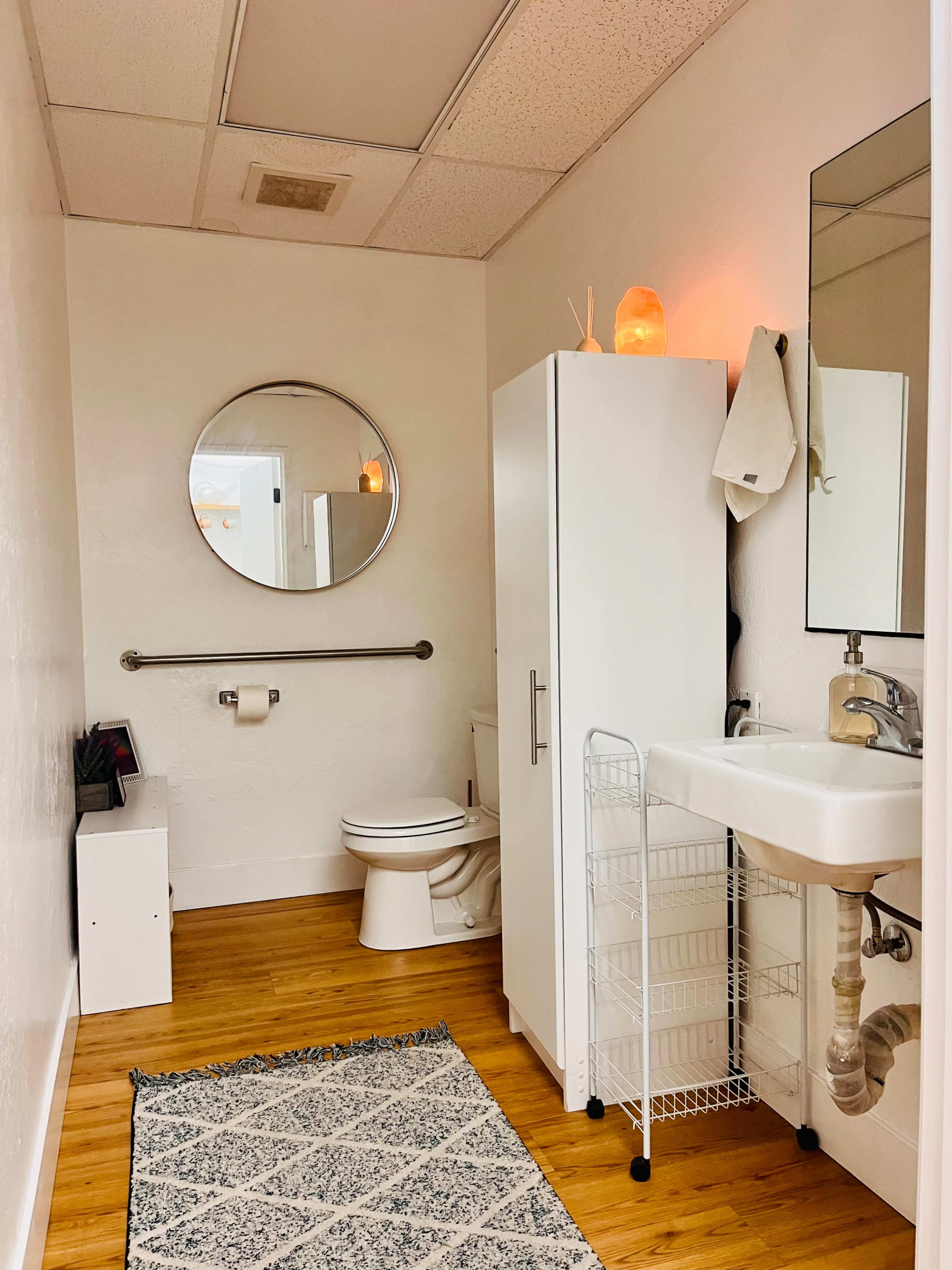 A clean and bright bathroom features a toilet, a white cabinet, a sink with a faucet, and a round mirror, all on a wooden floor with a patterned rug.