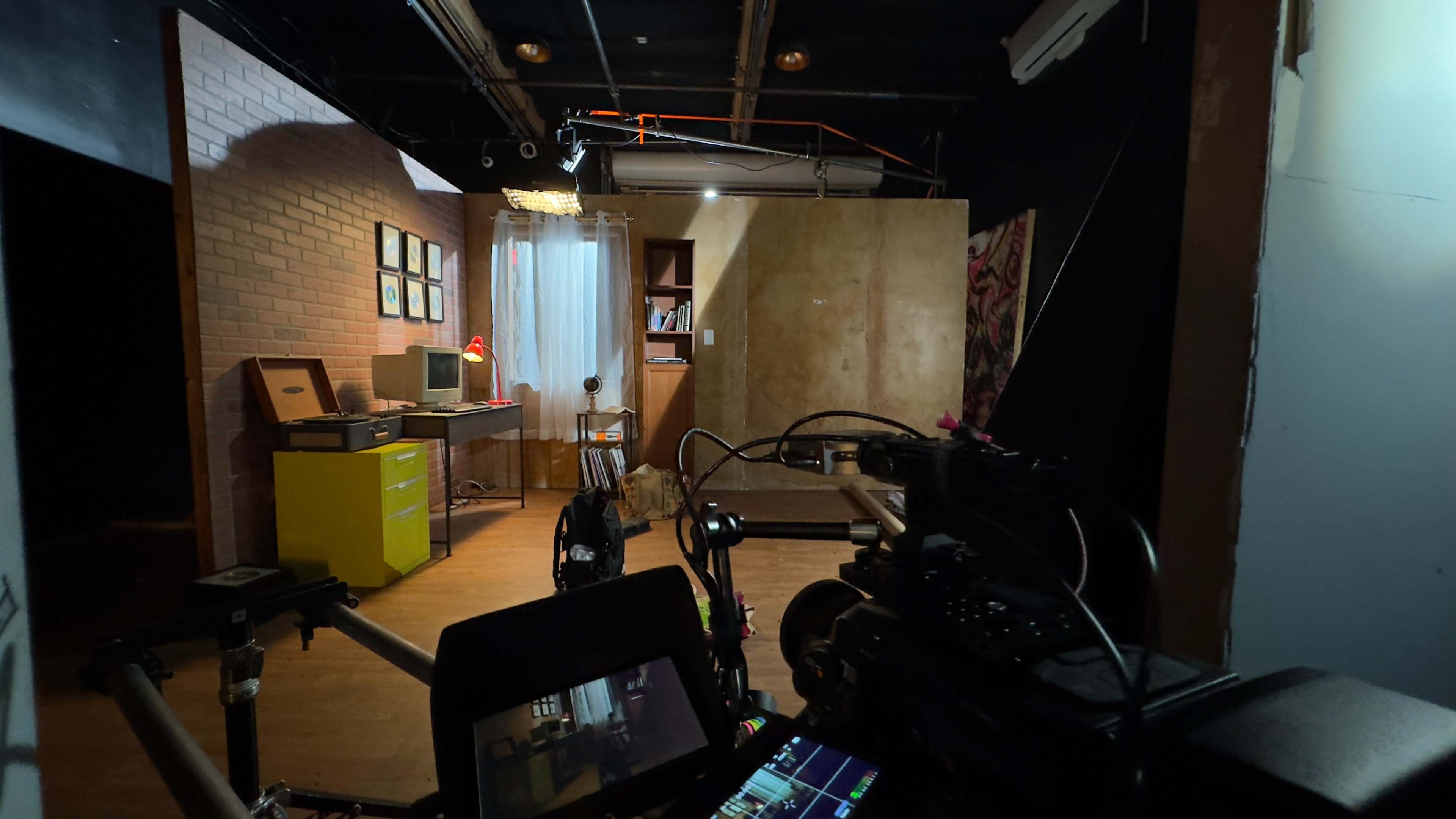 The image shows a film set featuring a small, well-lit room with a desk, a yellow cabinet, a shelf, and a window with sheer curtains.