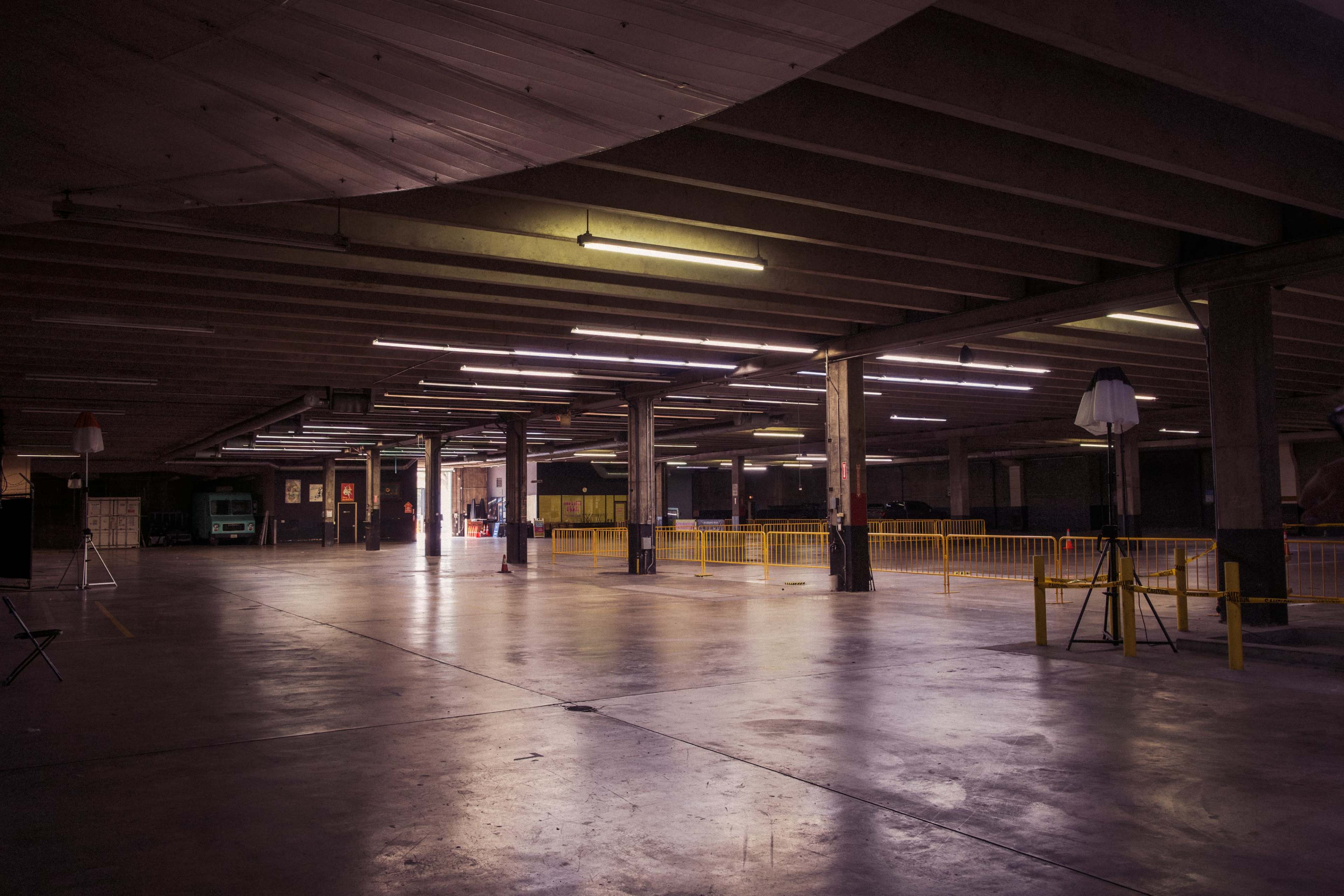 The image shows an empty indoor parking garage with fluorescent lights illuminating the concrete floor and yellow barriers marking off certain areas.