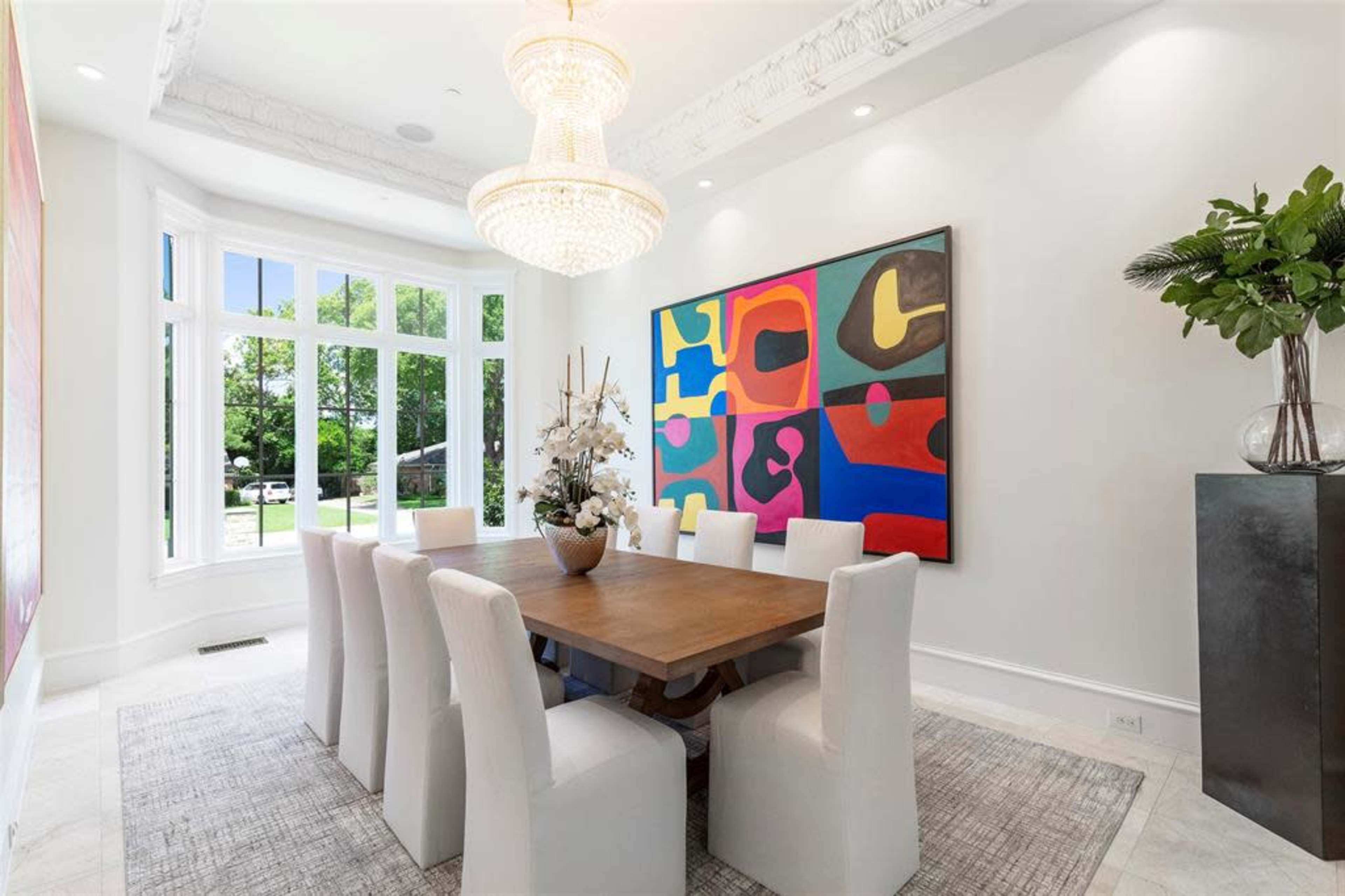 A modern dining room features a large wooden table surrounded by white upholstered chairs, a colorful abstract painting on the wall, and a chandelier overhead.