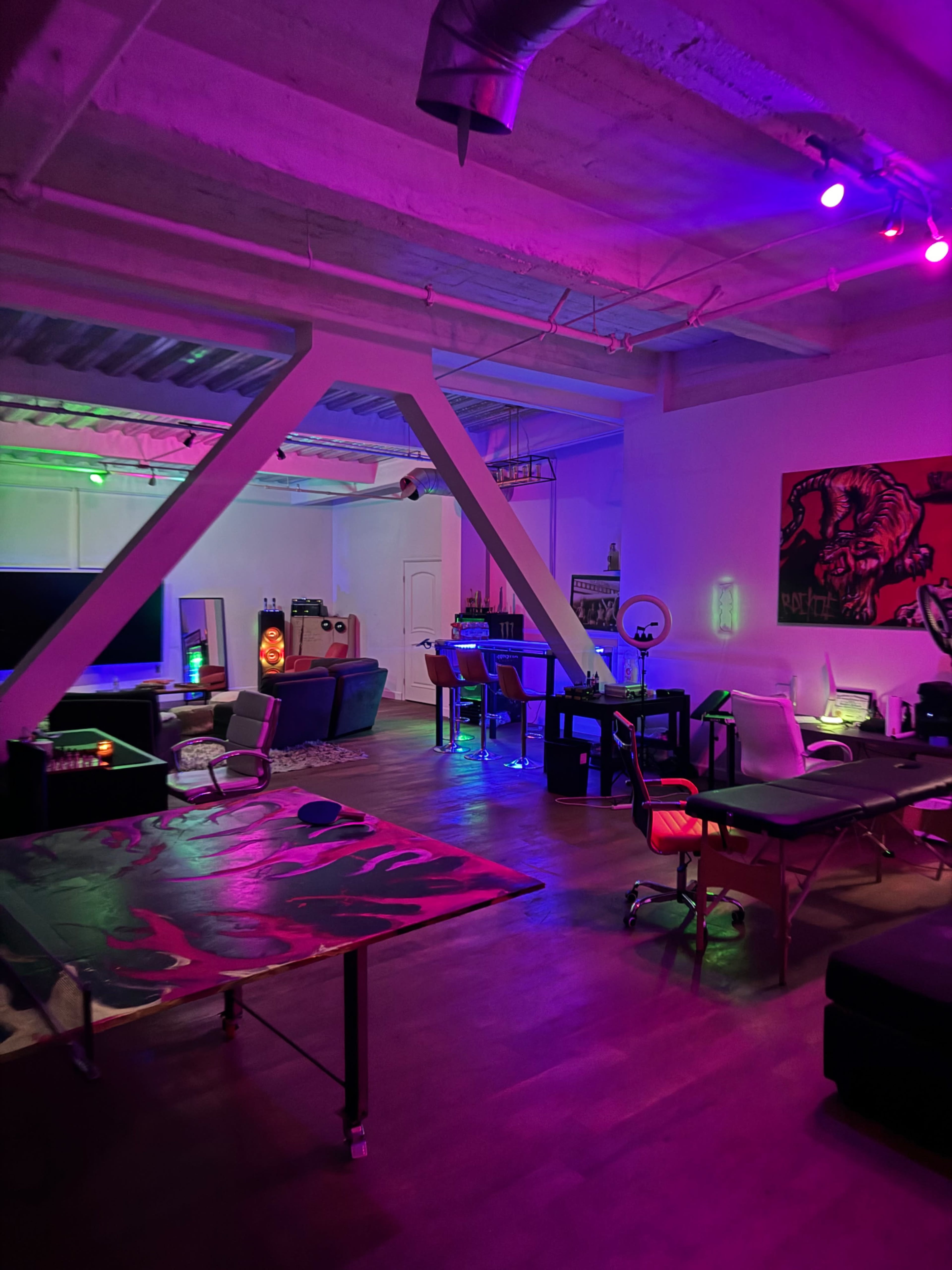 The image shows an artistic studio space with colorful lighting, featuring various furniture pieces including tables, chairs, and a large screen.