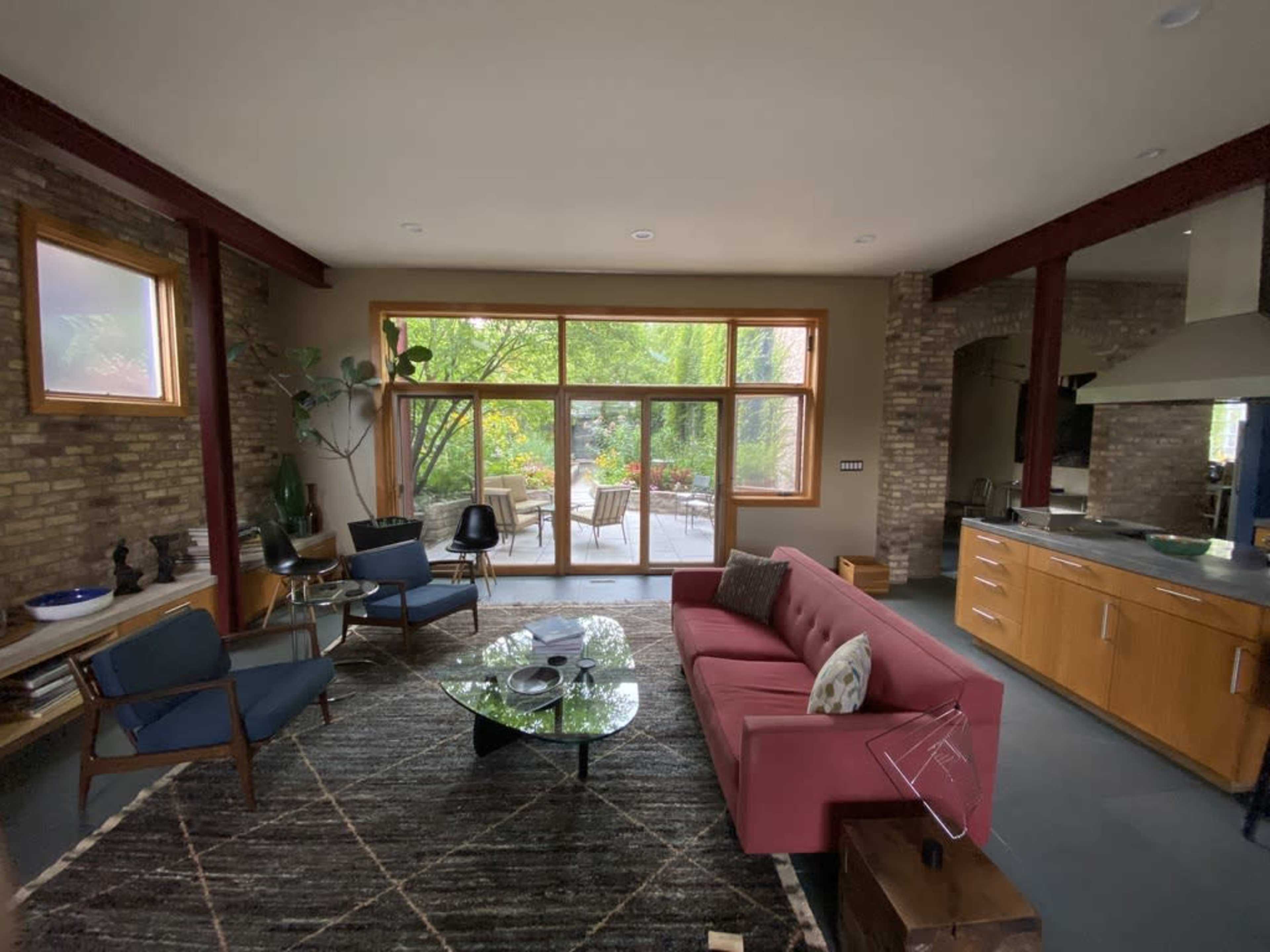 A modern living room with a red sofa, a glass coffee table, and large windows overlooking a green outdoor area.