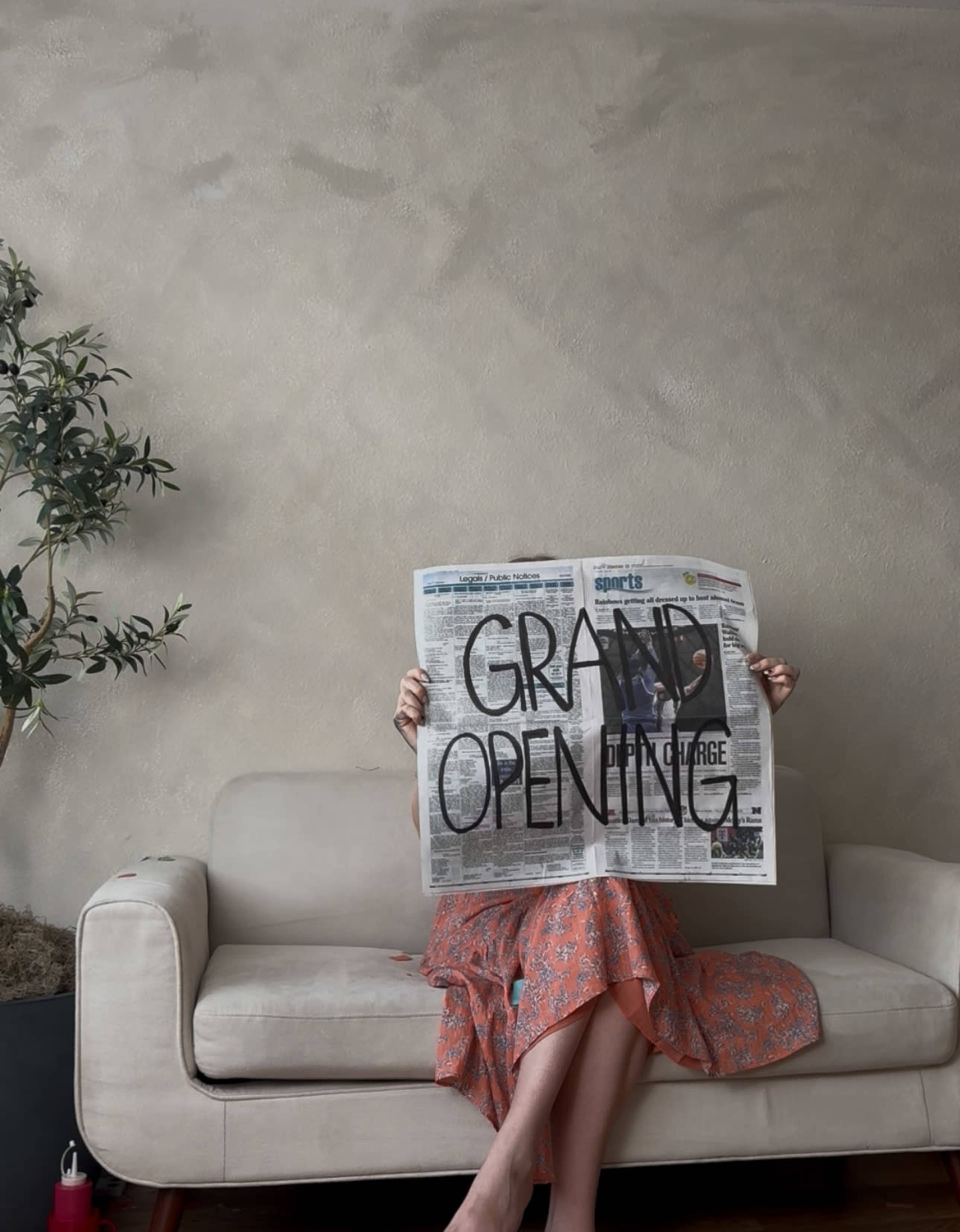 A person holding a newspaper with "GRAND OPENING" written in bold letters sits on a sofa, with a plant in the background.