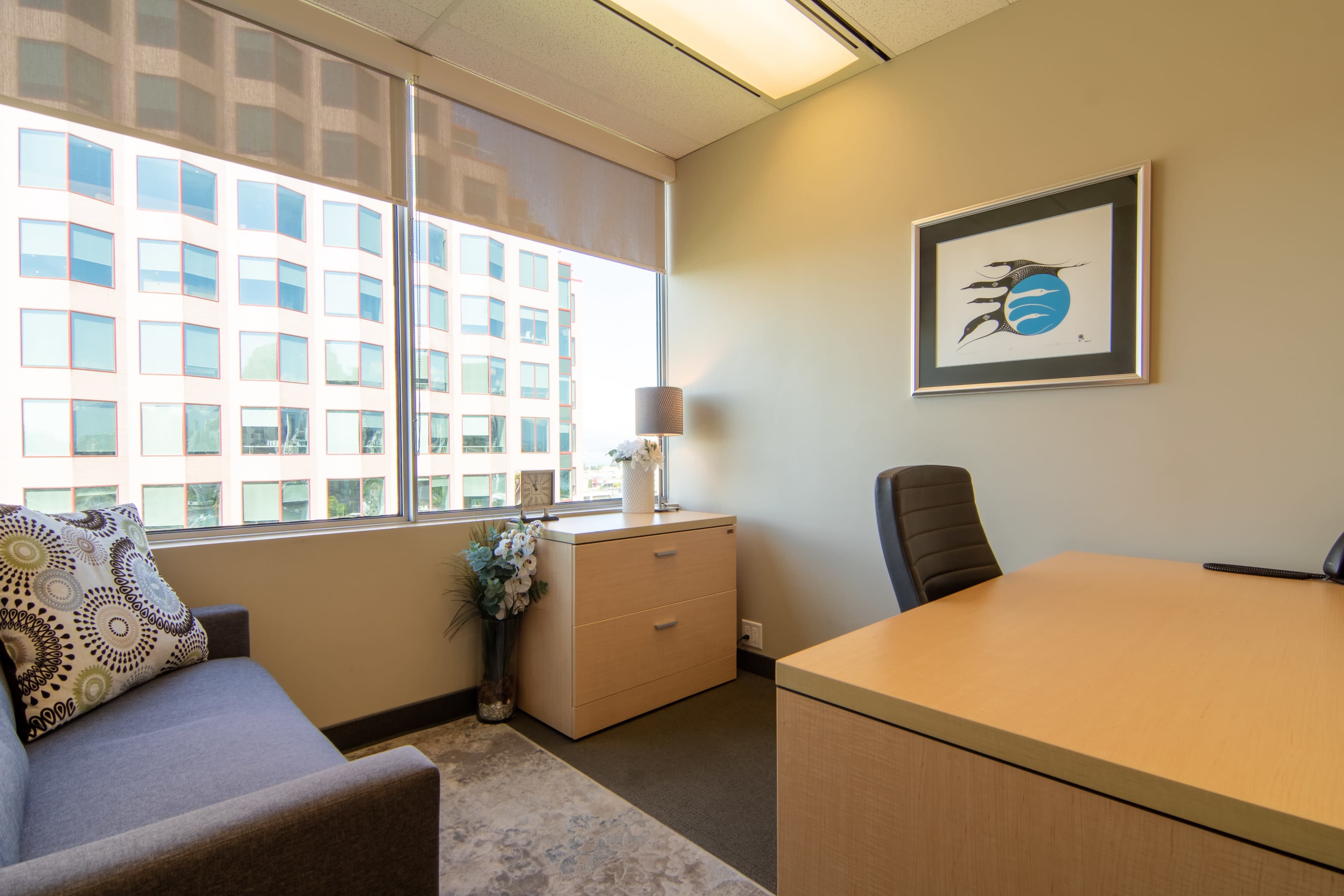 The image shows a modern office space with a sofa, desk, and large windows offering a view of a building outside.