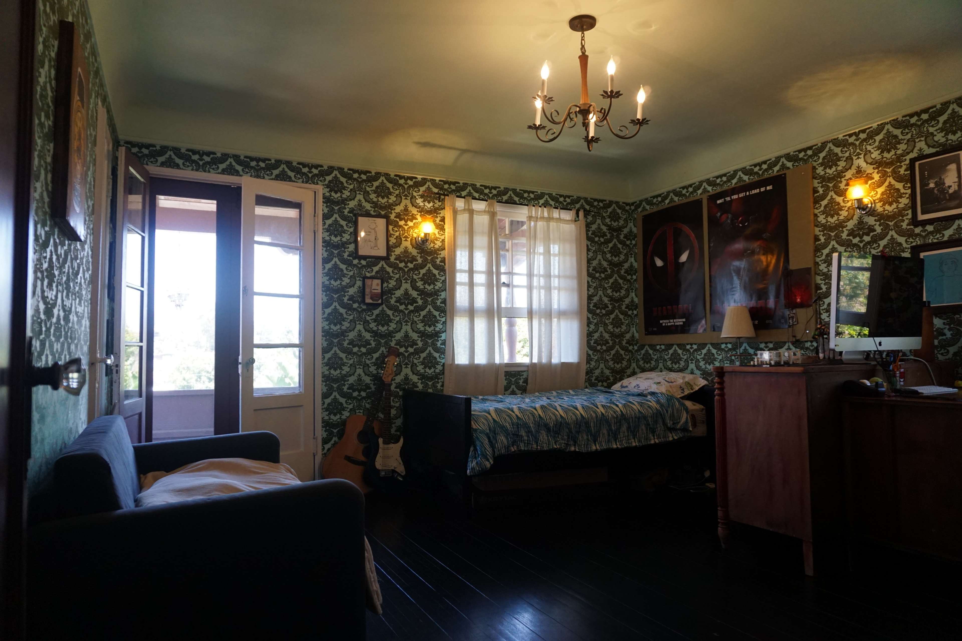 The image shows a bedroom with patterned wallpaper, a bed with blue bedding, a guitar in the corner, and a window letting in natural light.
