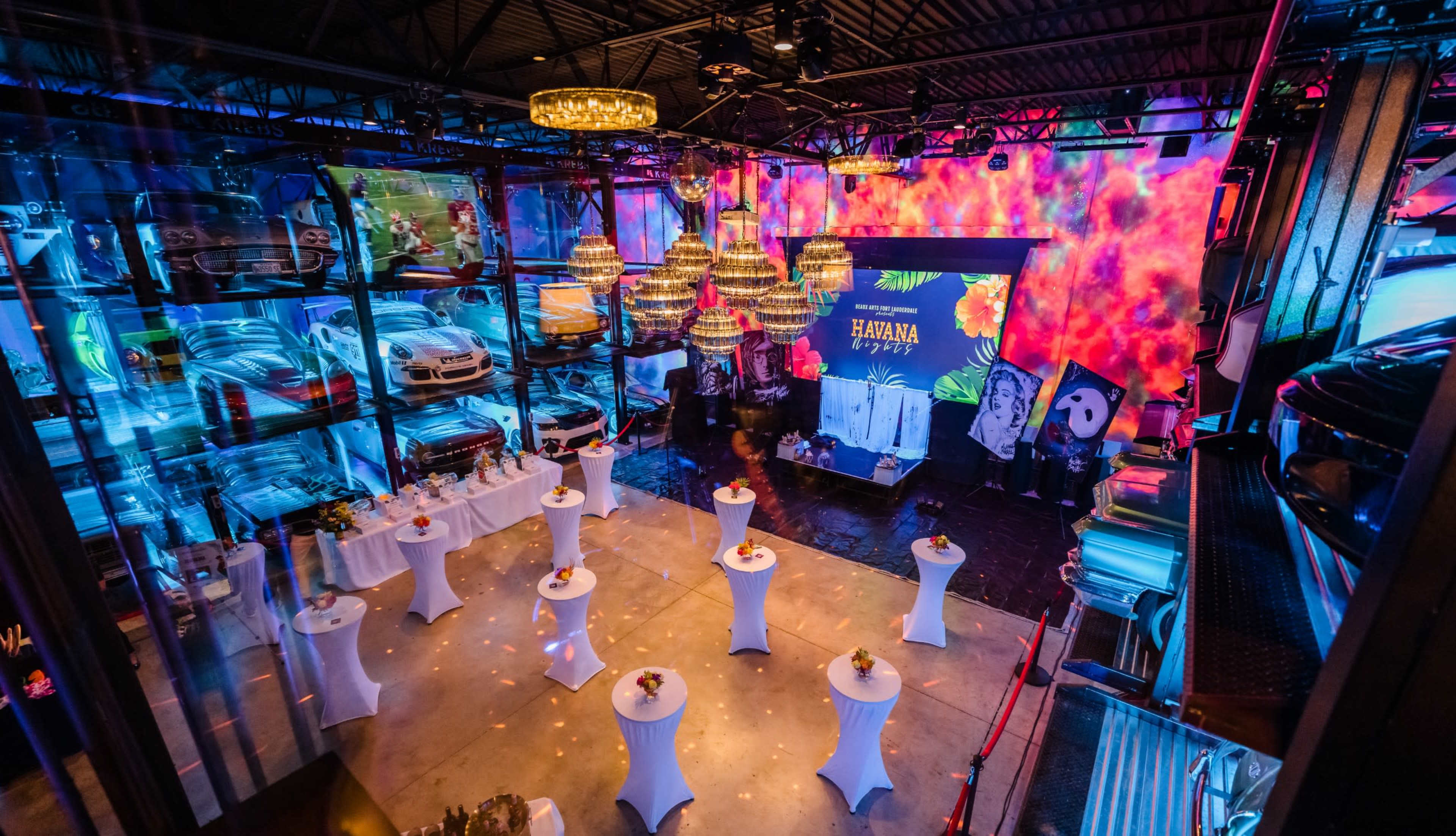 A spacious event venue featuring white cocktail tables arranged around a stage, with colorful lighting and car displays in the background.