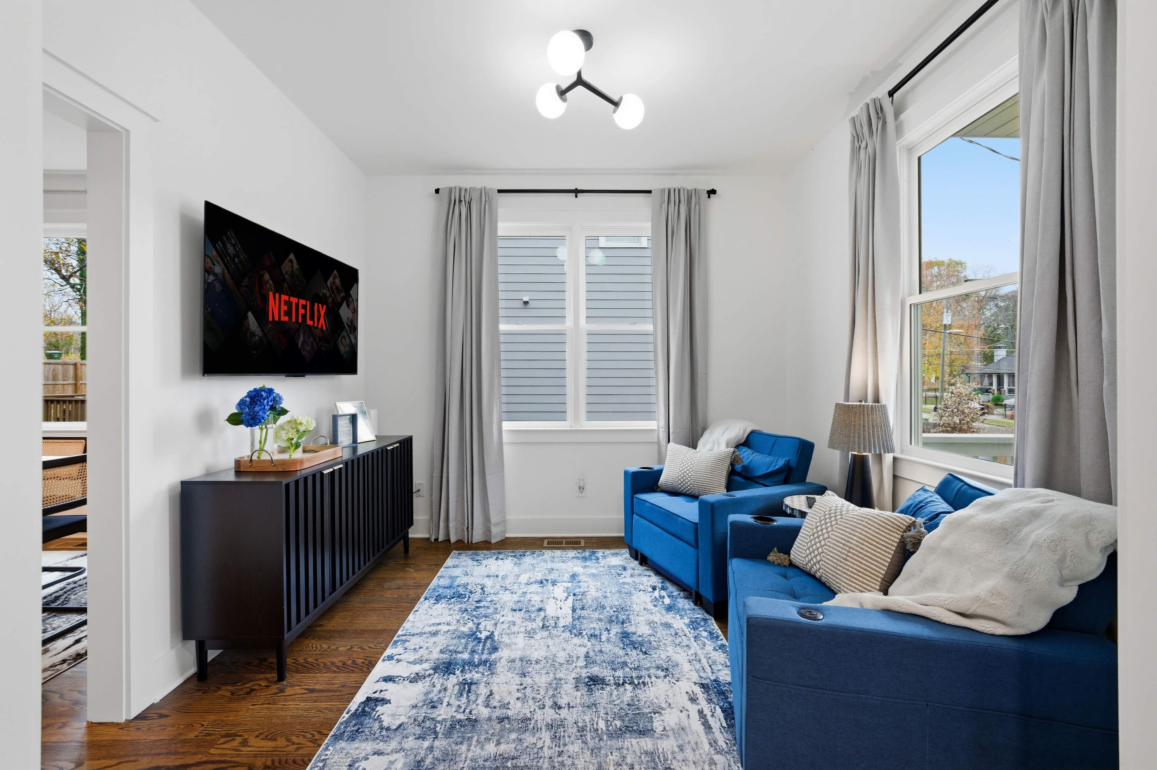 A bright living room features blue sofas, a modern light fixture, and a Netflix screen on the wall alongside large windows with light curtains.