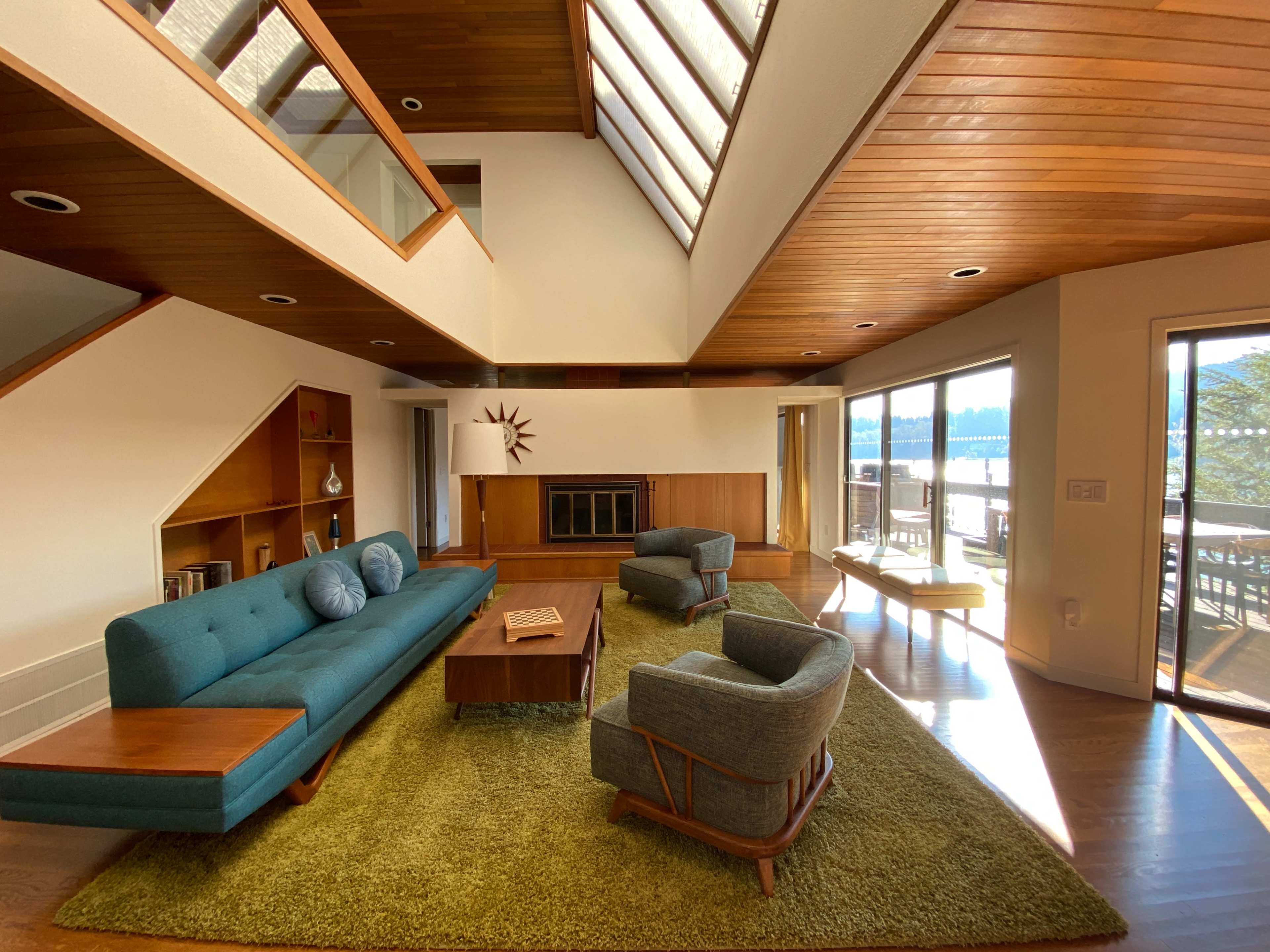 A modern living room featuring a turquoise sofa, two armchairs, a wooden coffee table, and large windows overlooking a deck and water view.