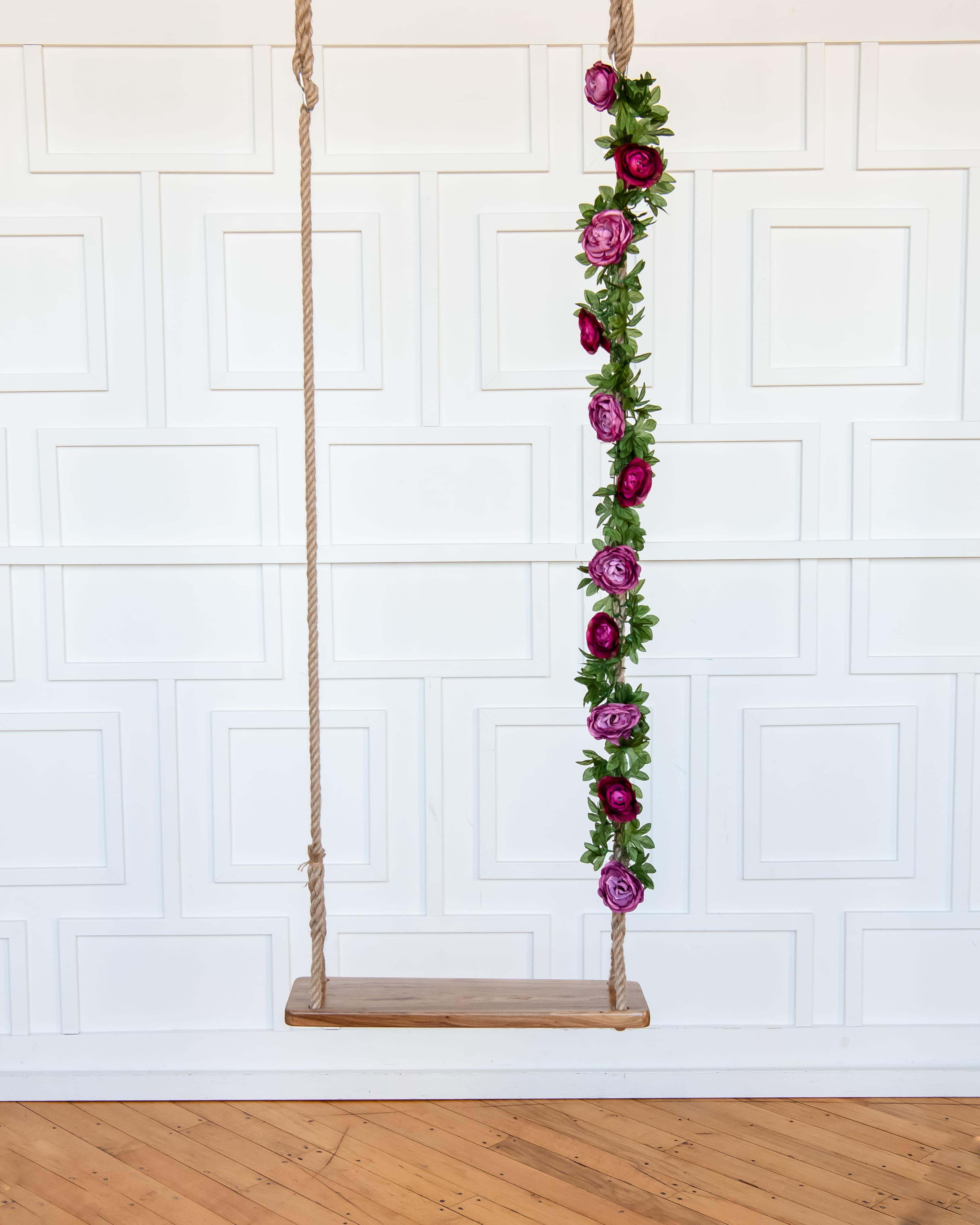 A wooden swing hangs from a white, paneled wall, adorned with a vertical arrangement of purple and pink flowers.