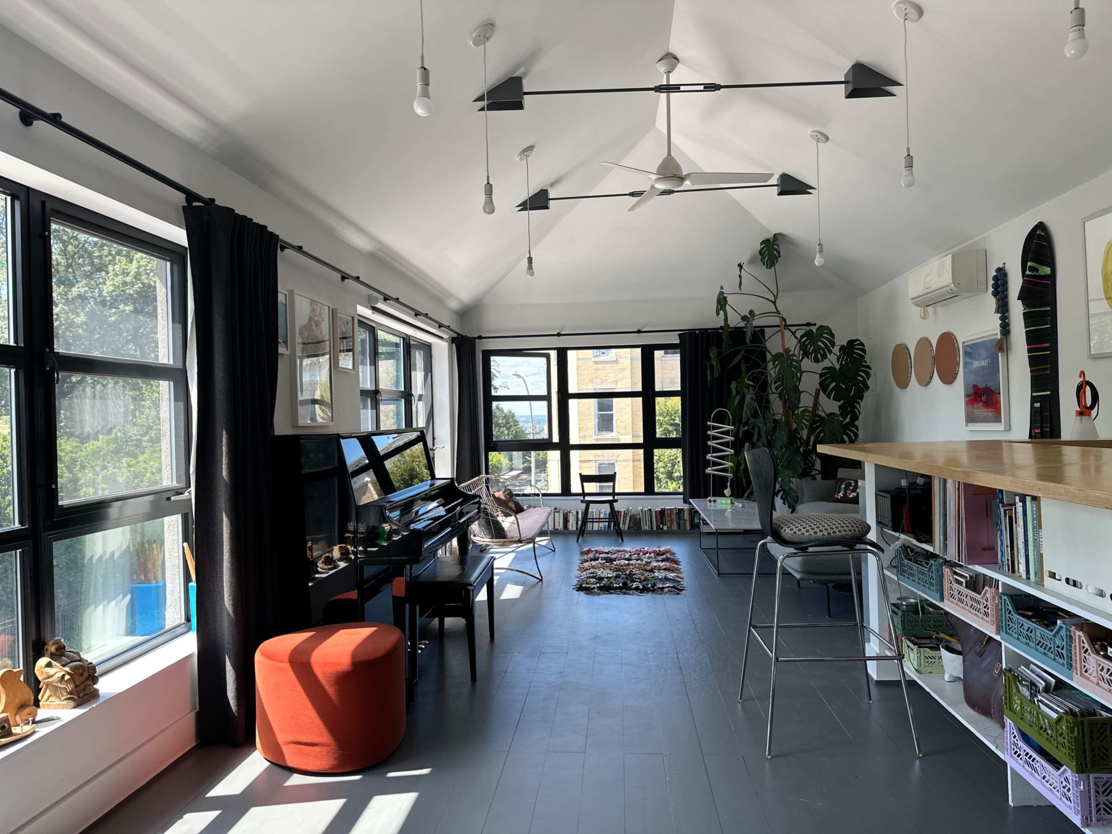 A sunlit room features large windows, a piano, a bar-height table, and various decorative elements including plants and artwork.