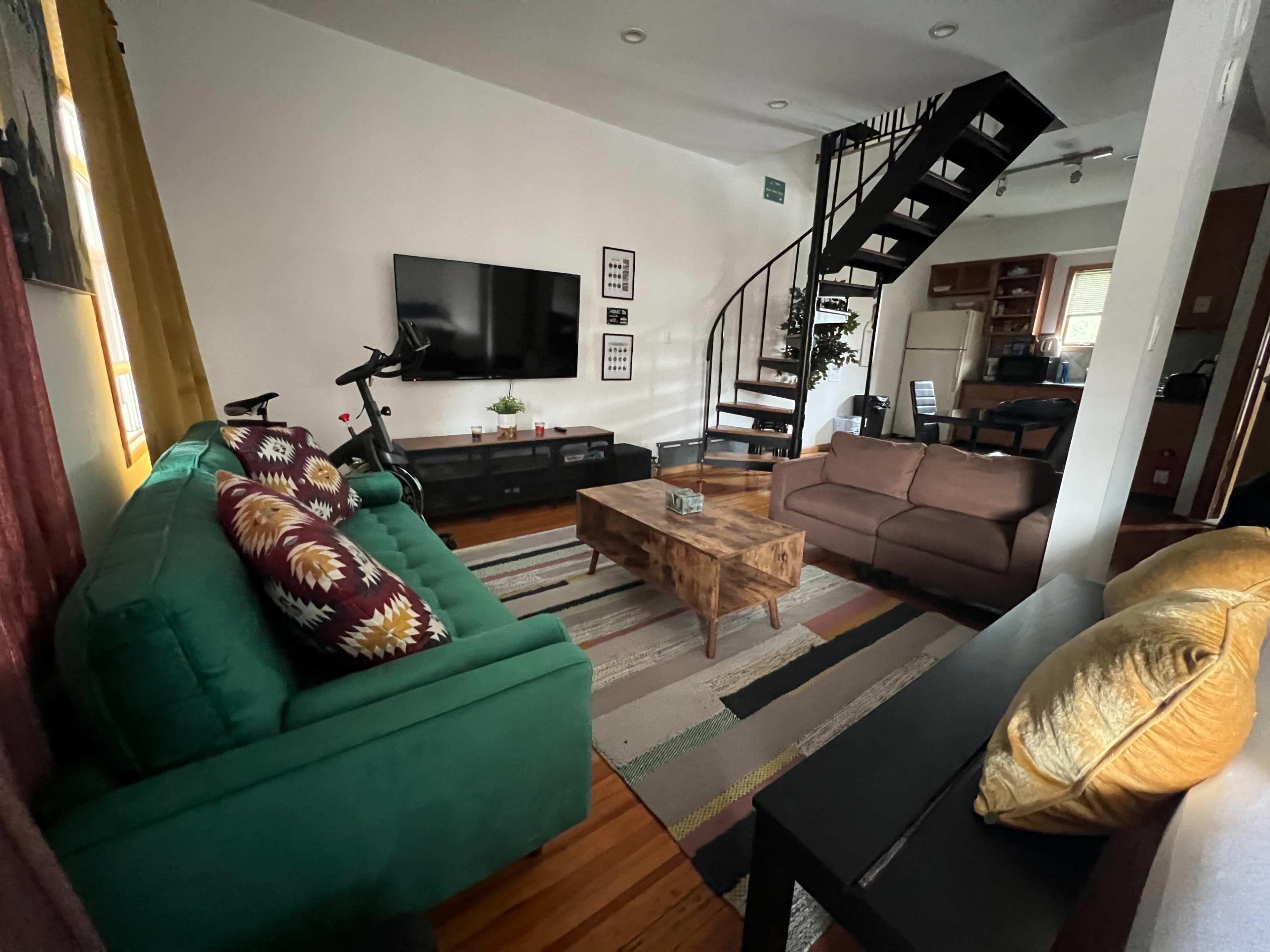 The image shows a living room with a green couch, a brown coffee table, a television mounted on the wall, a spiral staircase, and a view of a kitchen area in the background.