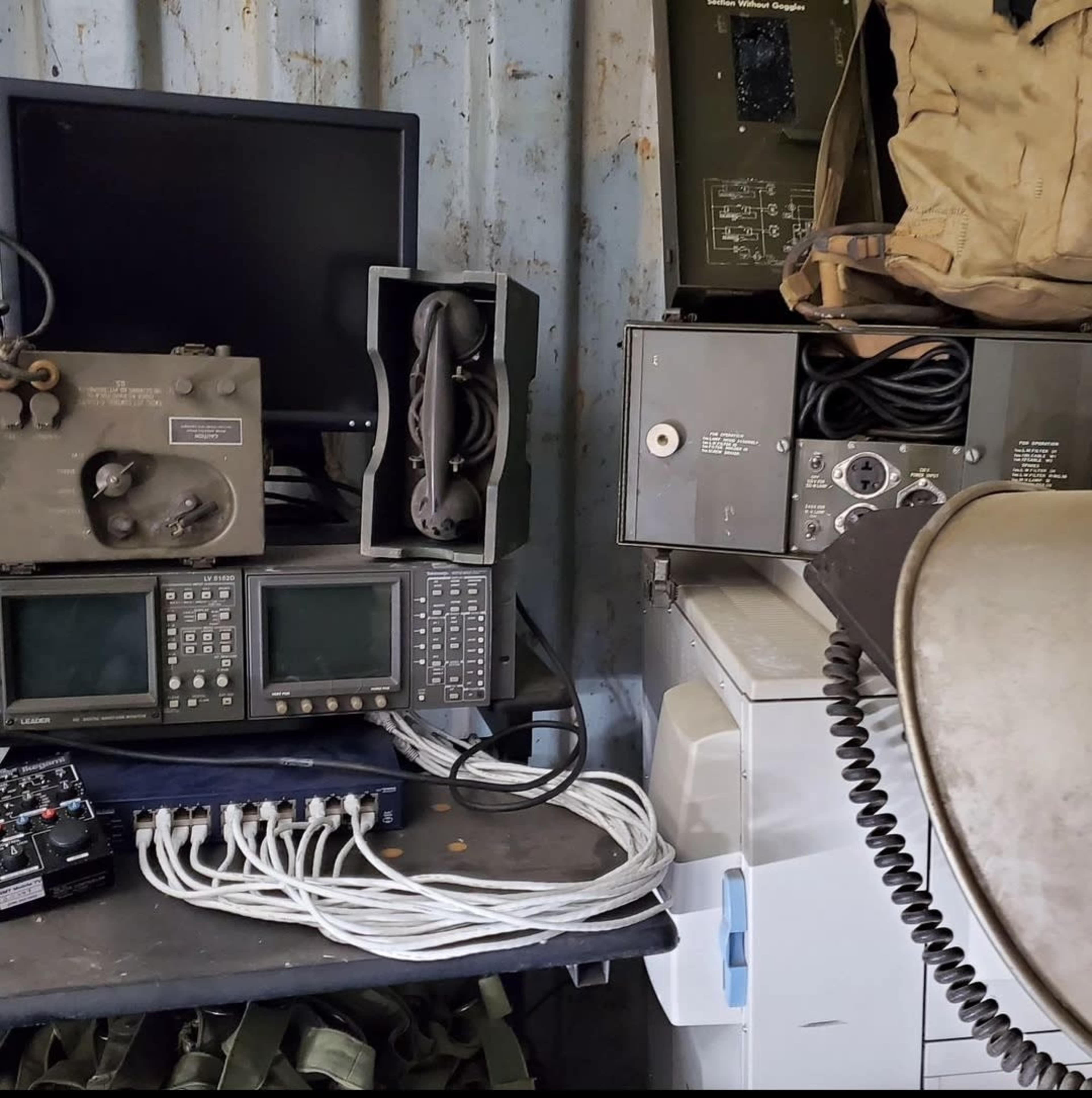 The image shows a cluttered workspace containing various pieces of communication equipment, including monitors, a printer, and numerous cables.