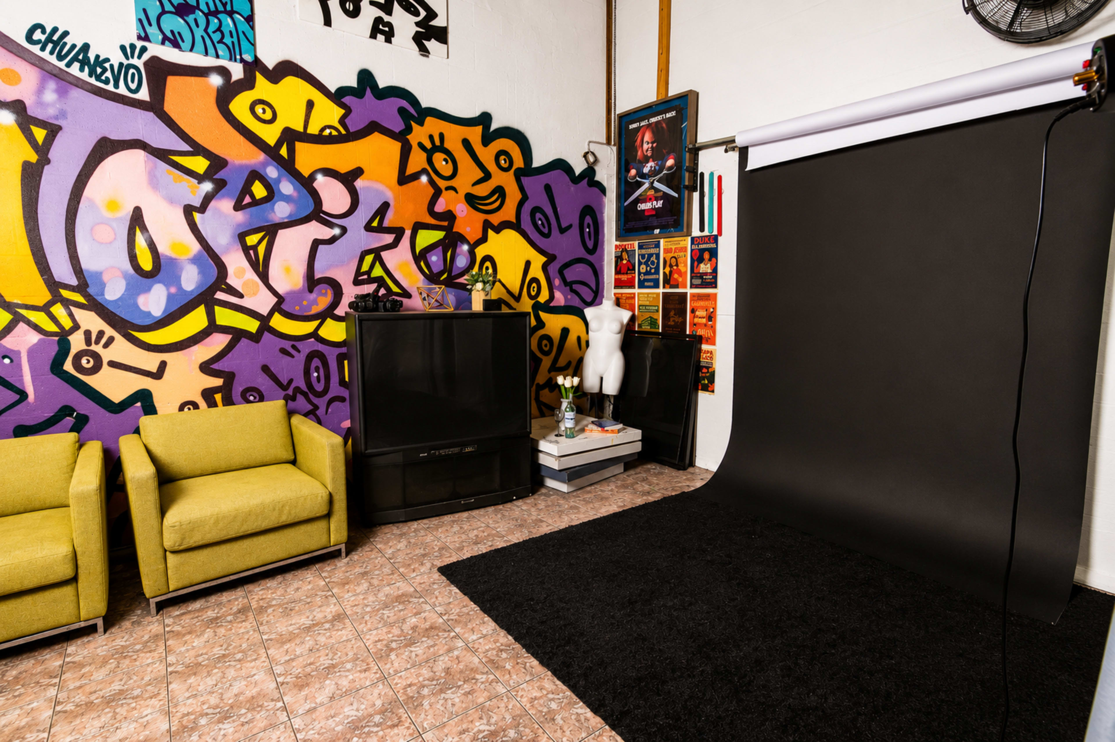 The image shows a vibrant studio space with a colorful graffiti wall, two green chairs, a black backdrop, and various decorations including posters and a mannequin.