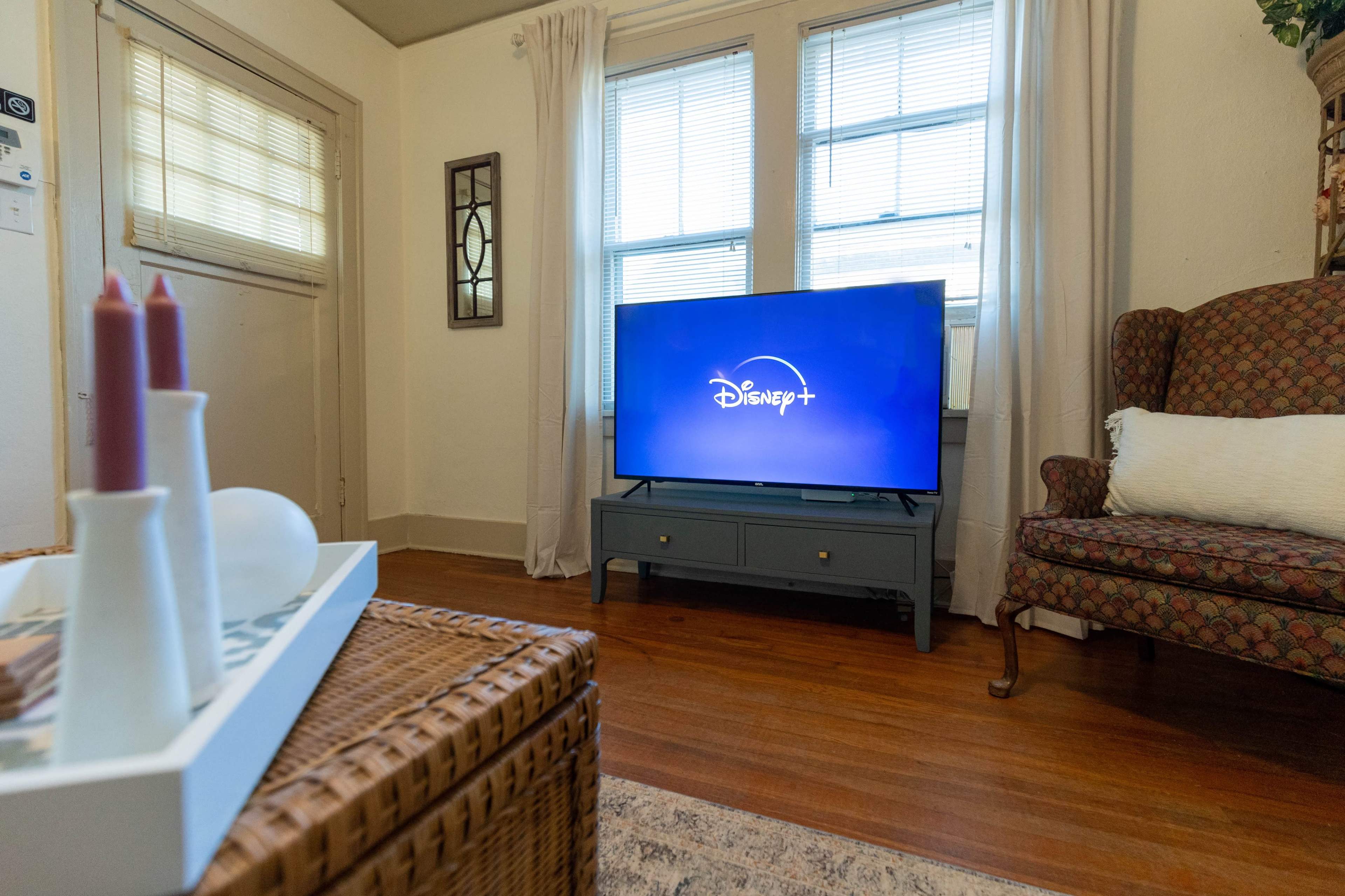 A living room with a television displaying the Disney+ logo, a wicker coffee table, and a comfortable armchair near a window.