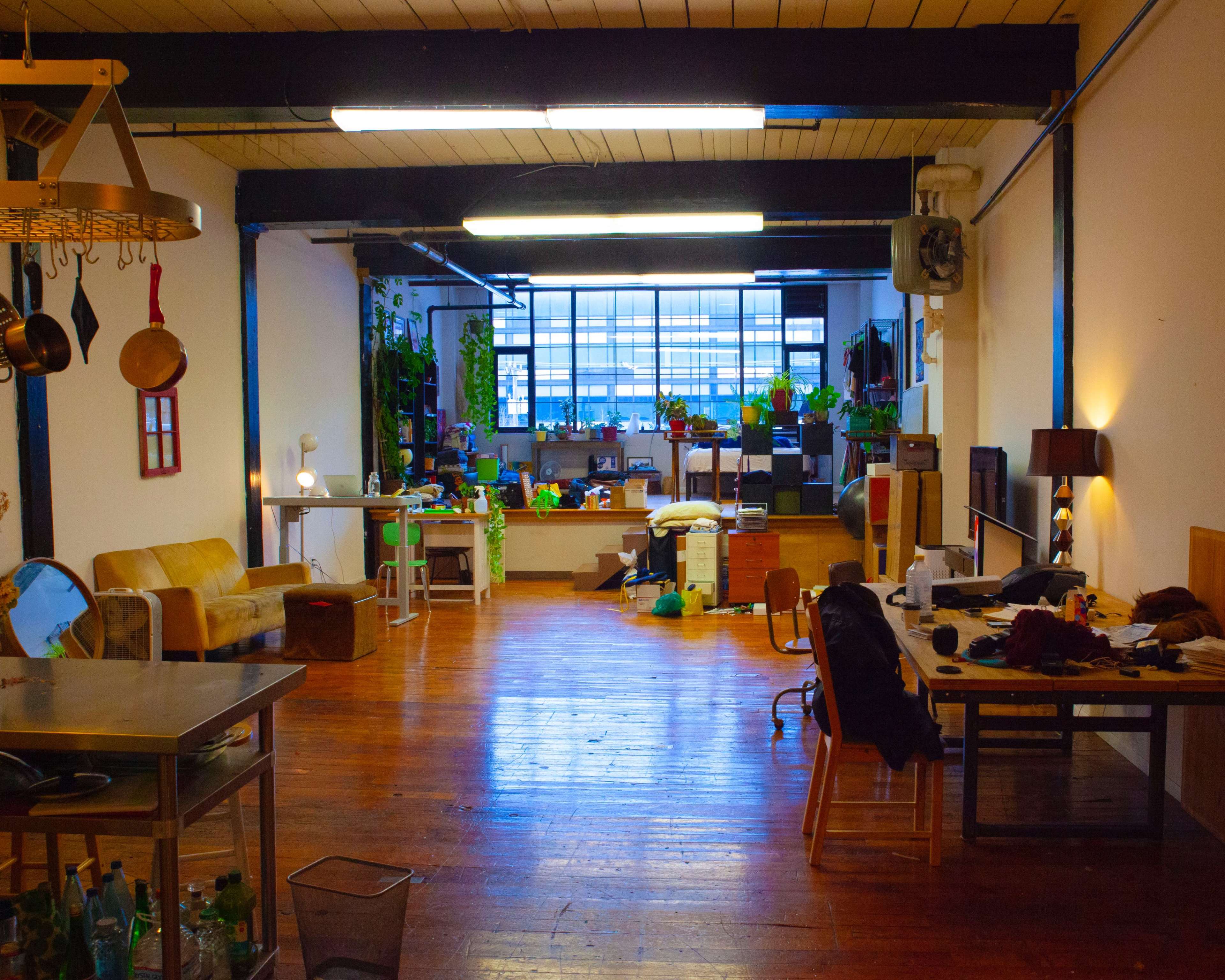 A spacious room with wooden floors, featuring various pieces of furniture, plants, and workstations scattered throughout.