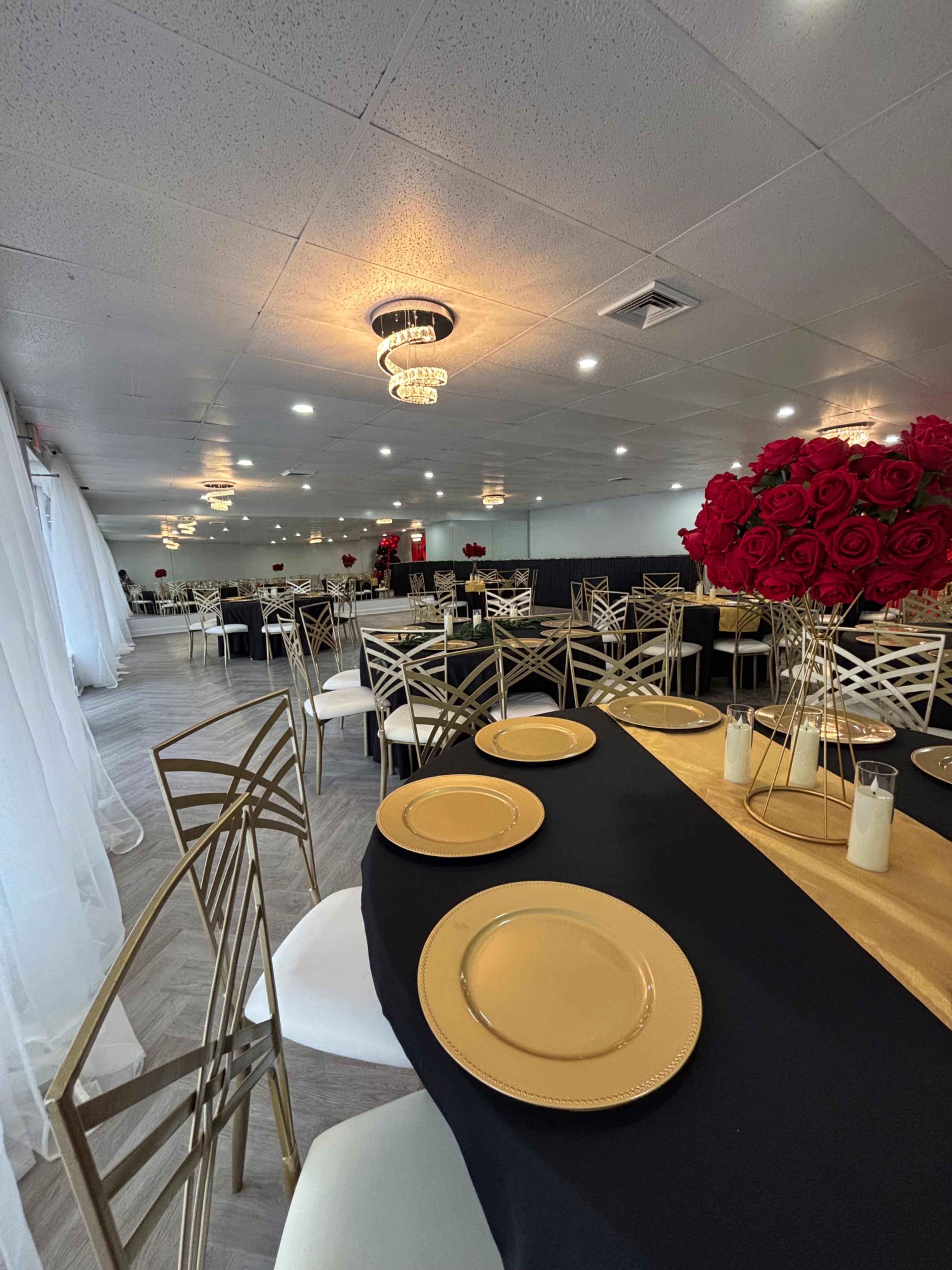 The image shows a elegantly arranged banquet hall with gold and black table settings and large floral centerpieces made of red roses.