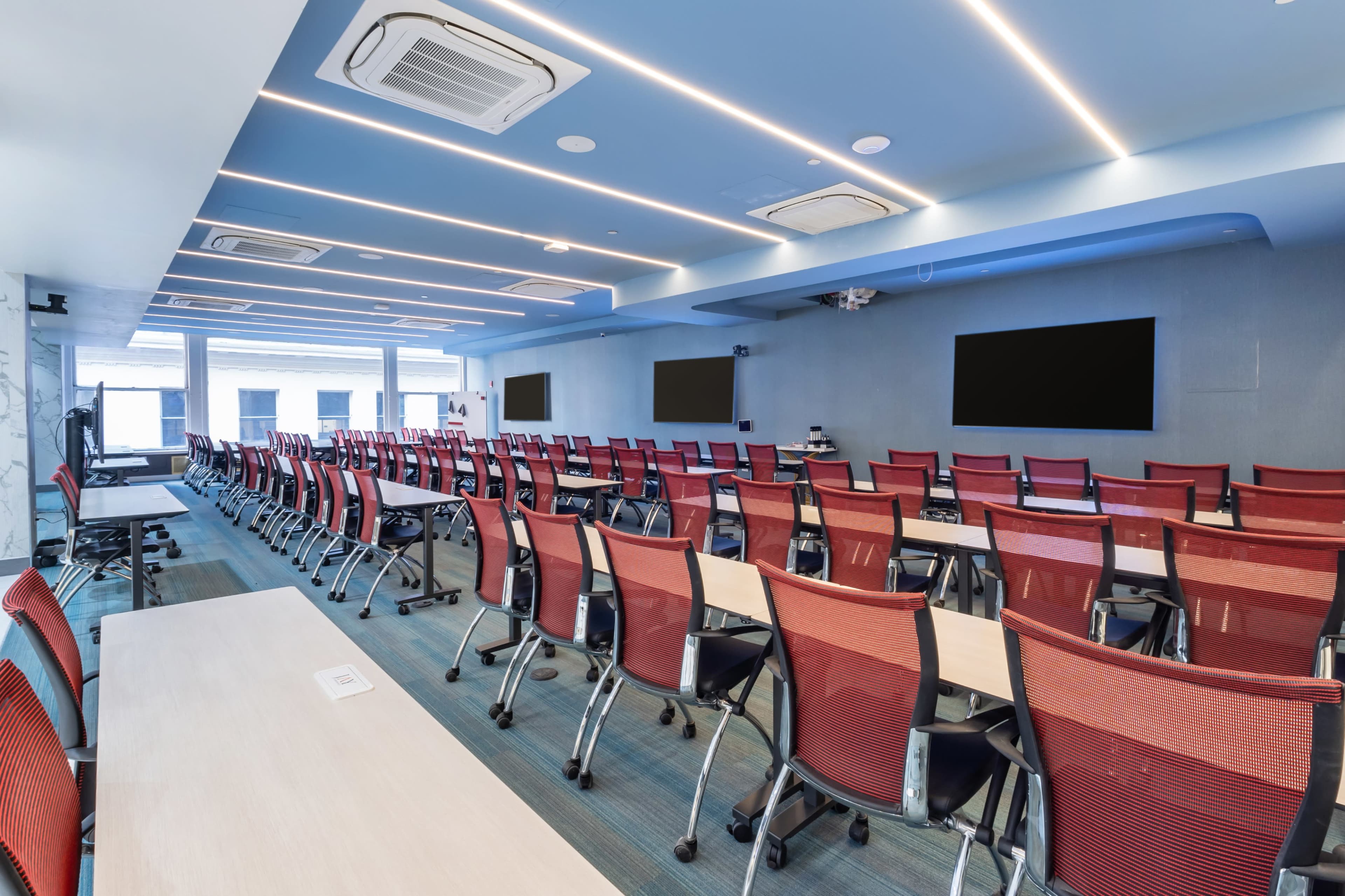 A modern conference room with rows of red chairs and tables facing multiple large screens.