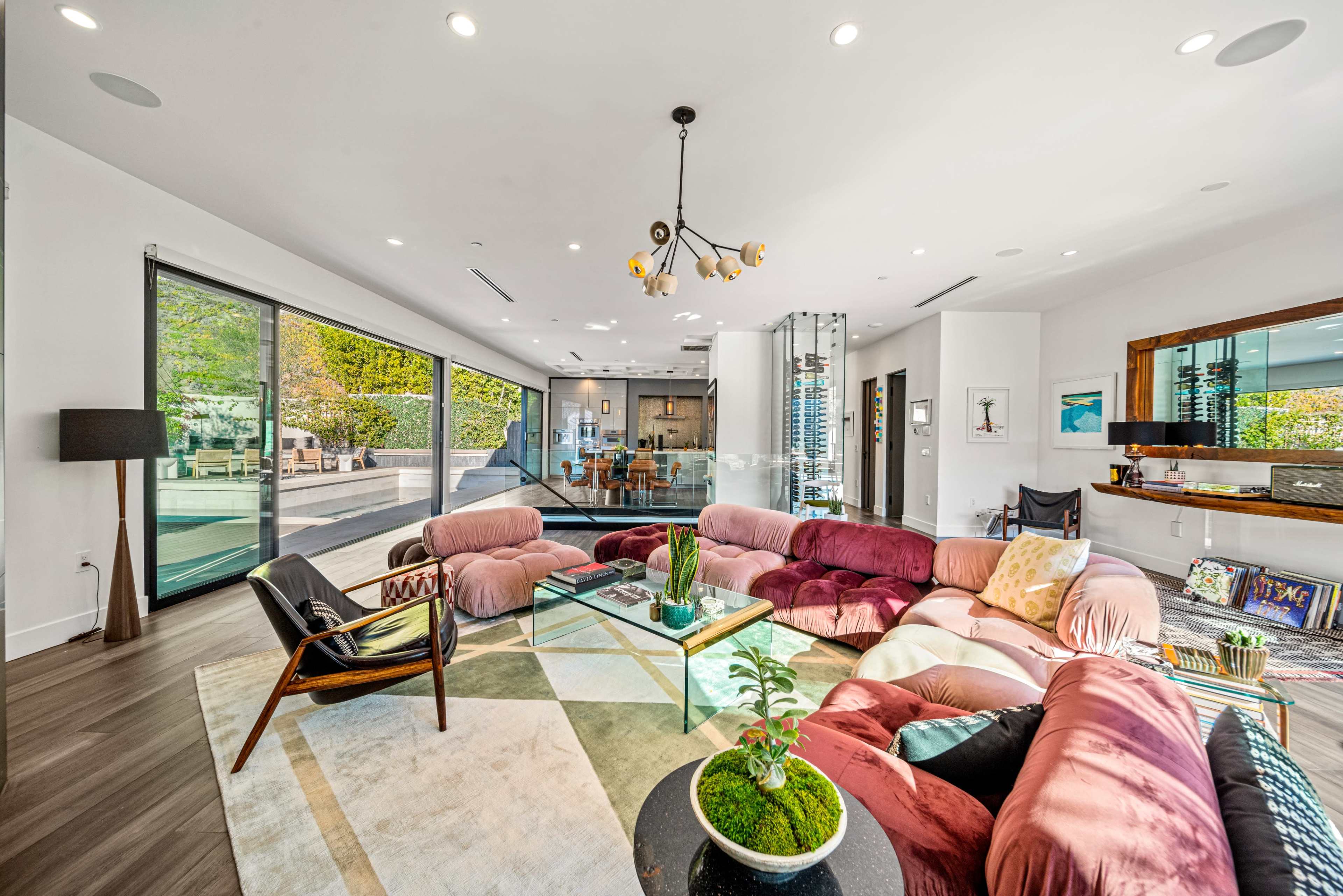 The living room features a large, curved sectional sofa, a glass coffee table, and expansive windows overlooking a landscaped outdoor area.