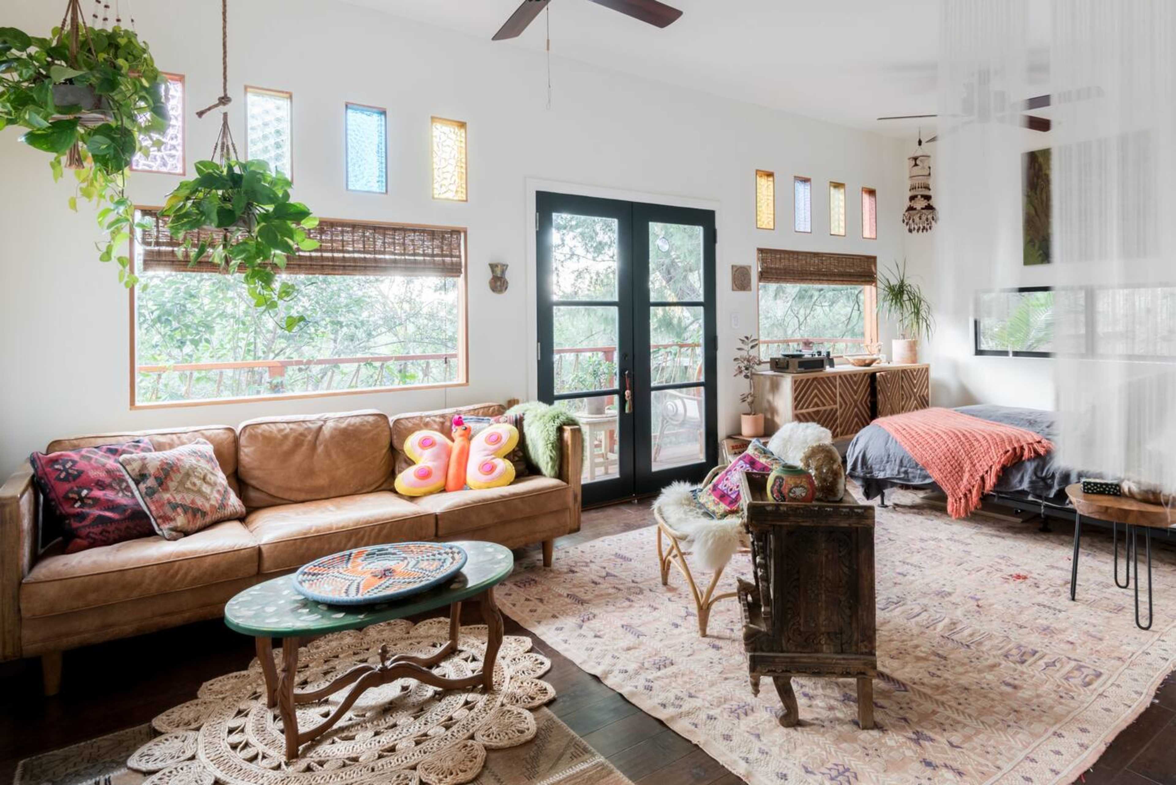 A cozy living room furnished with a leather sofa, colorful pillows, a circular table, and decorated with plants and natural light filtering through patterned windows.