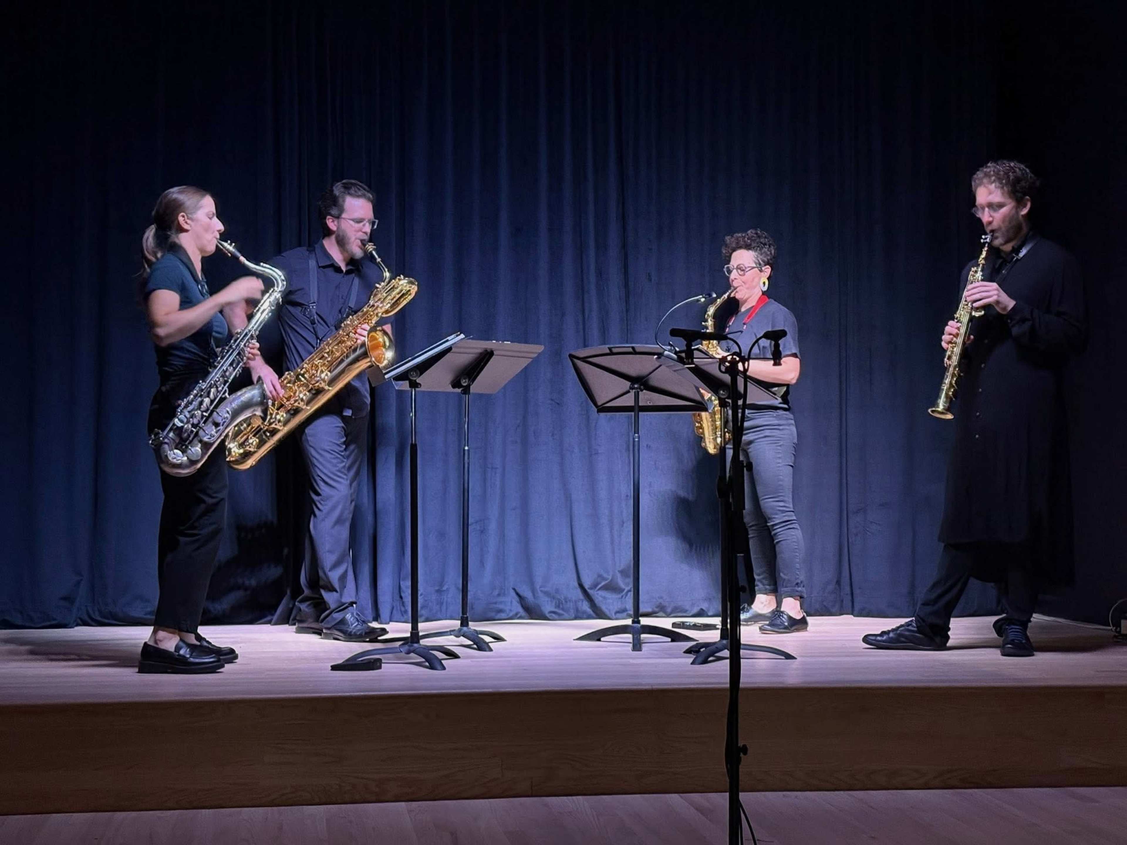 Four musicians play saxophones on stage, standing in front of a dark curtain.
