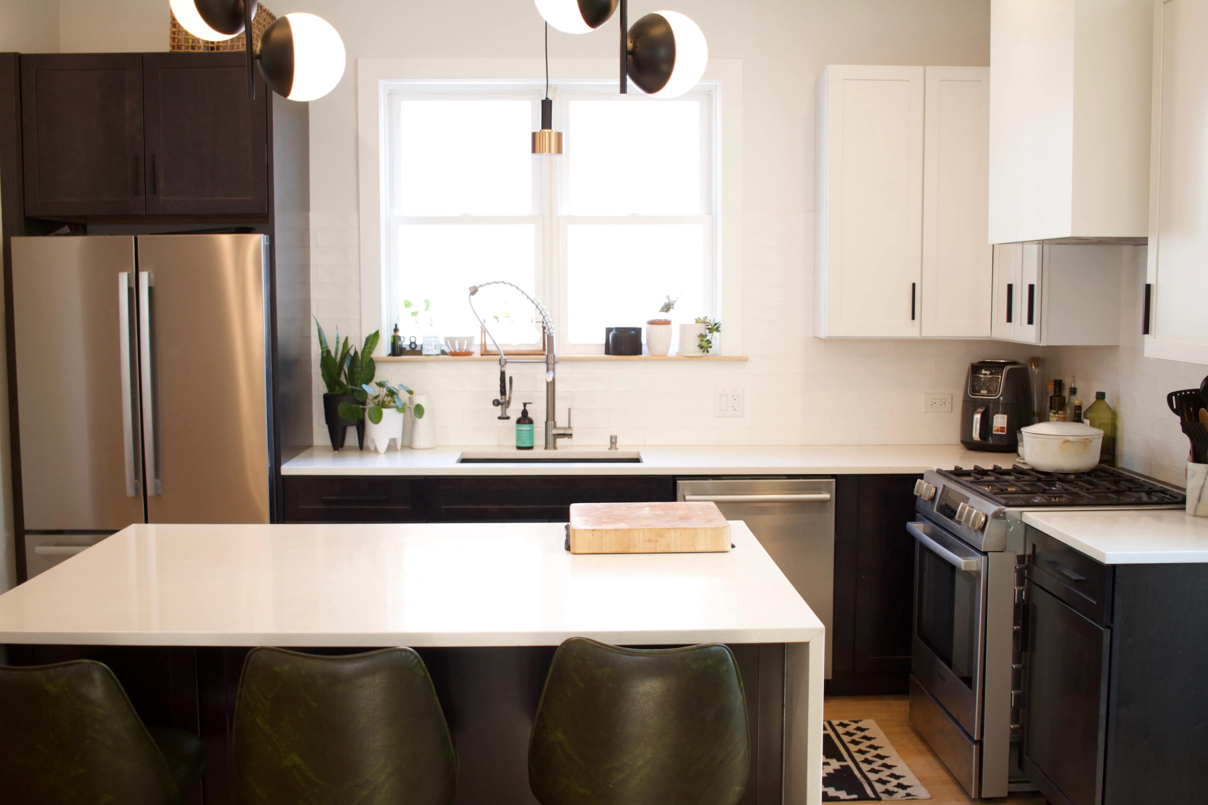 A modern kitchen featuring a central island with three green chairs, stainless steel appliances, a double sink, and a window with plants.
